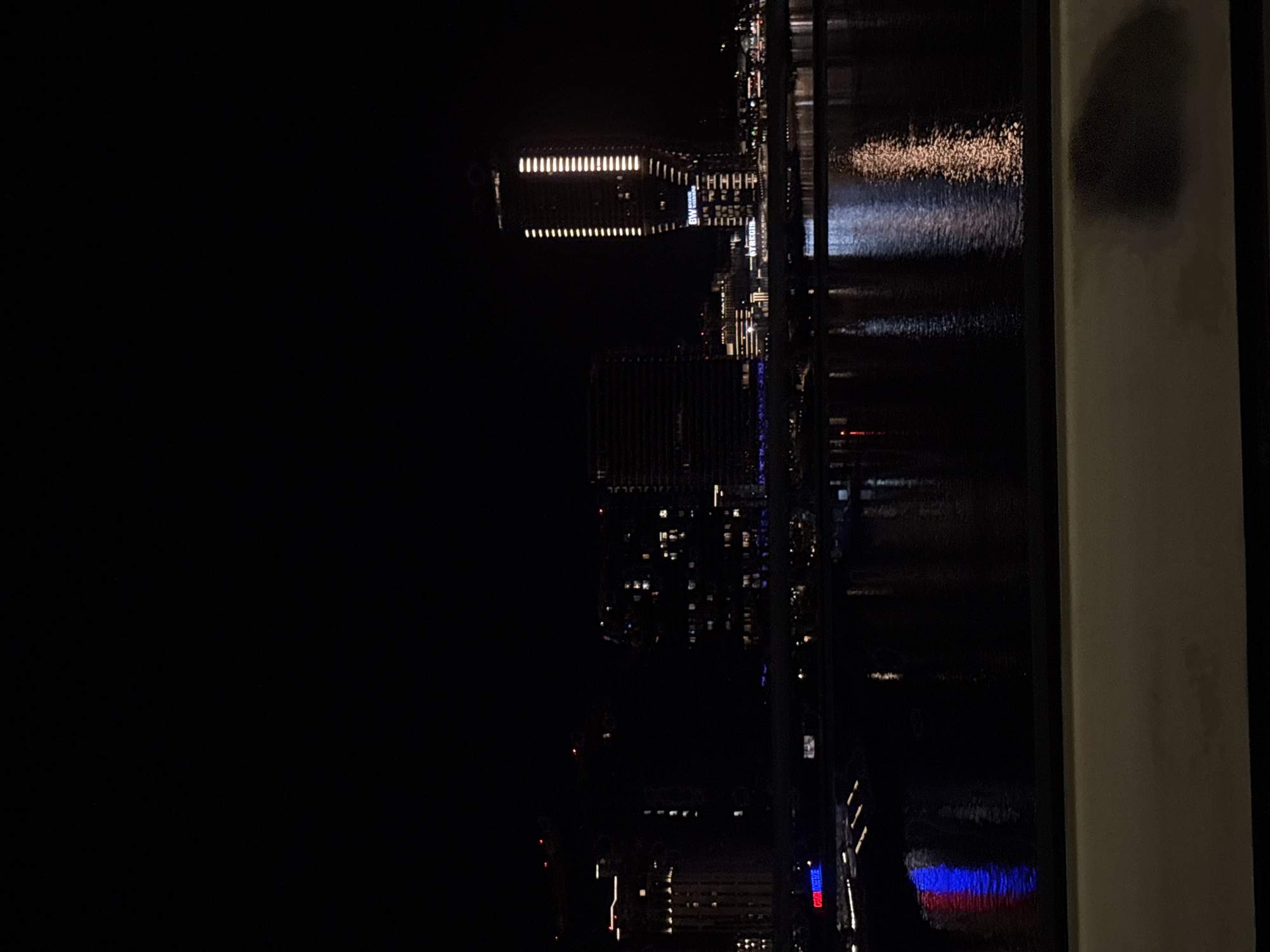 The image depicts a nighttime view of Belgrade, Serbia, from a high vantage point, likely a window. The scene includes illuminated buildings reflecting in a body of water. Specifically, the reflections show blue and red lights. The right edge of the frame includes a partially visible wall or structure, suggesting the view is framed by an interior space. The rest of the scene is dark.