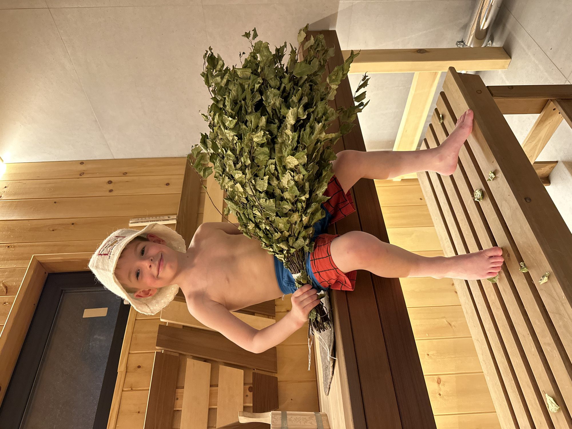 A young boy in a white sauna hat and red-and-blue swim trunks lies on wooden benches in a sauna. He is holding a bundle of birch branches. The image was taken indoors in Прикамский, Russia.
