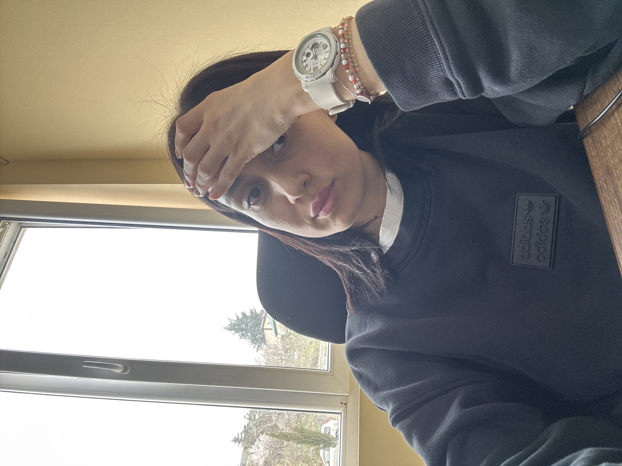 A female individual is depicted from the chest up, seated indoors. She has dark hair and a neutral expression with slightly parted lips. Her right hand is positioned on her forehead, fingers lightly spread. She is wearing a dark-colored, likely black or dark grey, long-sleeved sweatshirt with a black embossed Adidas logo patch on the left chest, over a white undershirt. Her left wrist is adorned with a white BABY-G brand watch and two distinct beaded bracelets, one primarily orange and red, the other with lighter, possibly pink beads and a small silver charm.
Behind the individual, a large window with white frames reveals an overcast sky and an outdoor urban landscape. The exterior view includes a hillside populated with various trees, notably a tall, slender coniferous tree and a deciduous tree with white blossoms, indicating spring. A light-colored vehicle, possibly a white car, is partially visible on a street or driveway below the hillside. Further up the slope, a building with a light-colored facade and a dark roof is visible among the vegetation. The indoor environment features a plain light brown-yellow wall and the dark-colored backrest of an office chair. This hilly urban setting with specific vegetation and architectural style is consistent with the city of Tbilisi, Georgia.