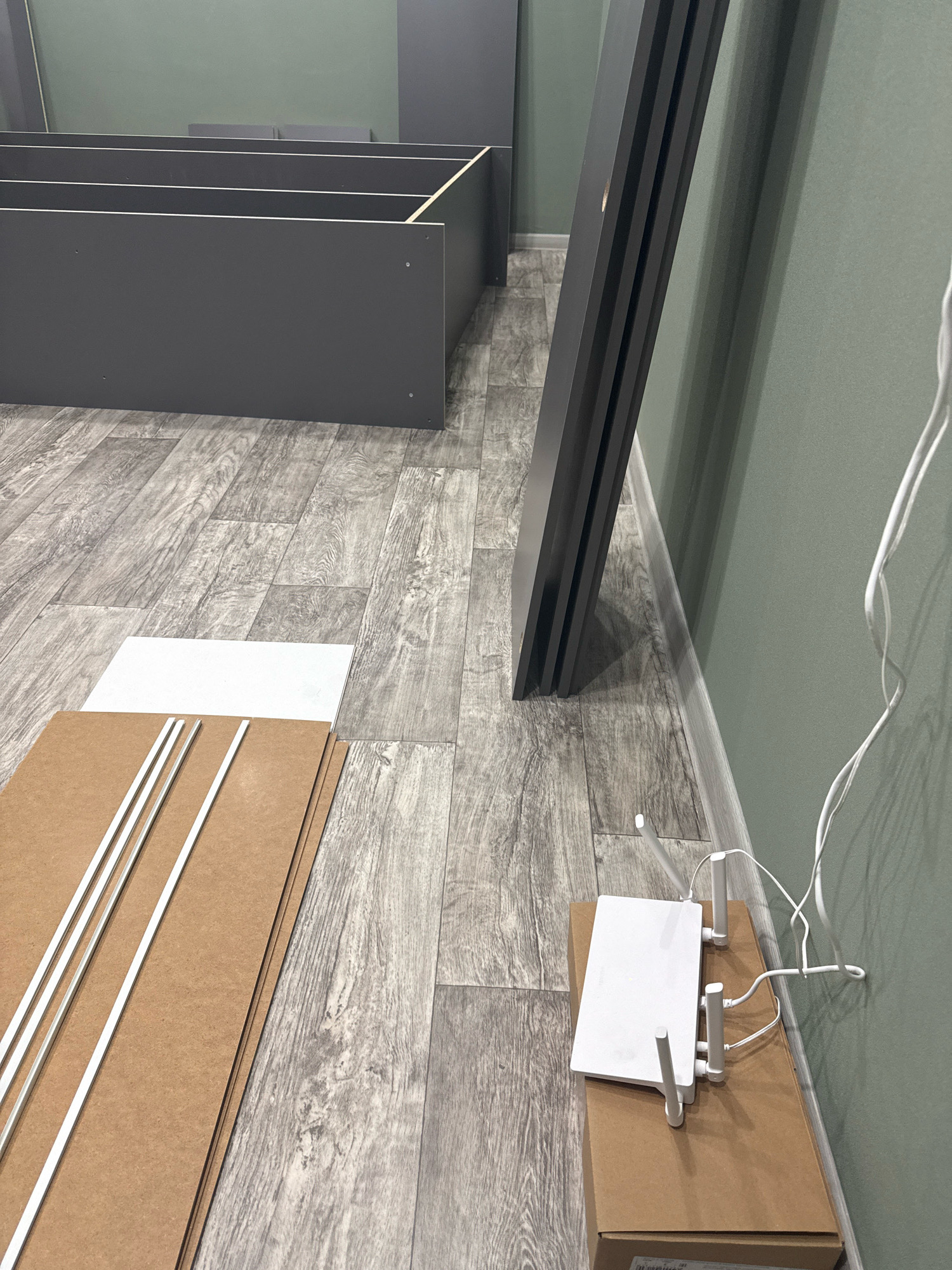 The image depicts an indoor scene, likely a room undergoing assembly or renovation. In the foreground, there are several large, flat pieces of particleboard or MDF, some with white trim pieces laid on top of them. To the right, on top of a cardboard box, sits a white wireless router with several antennas. White wires are connected to the router and trail off the right side of the frame. In the background, partially assembled dark gray shelving units are visible, along with a section of a dark gray vertical support beam. The floor is covered with laminate flooring that mimics the appearance of distressed wood planks. The walls are painted a muted green. There are no people visible in the image. The scene suggests a domestic or commercial space where furniture assembly is in progress. There is no specific visual information to link this image to Cheboksary, Russia.