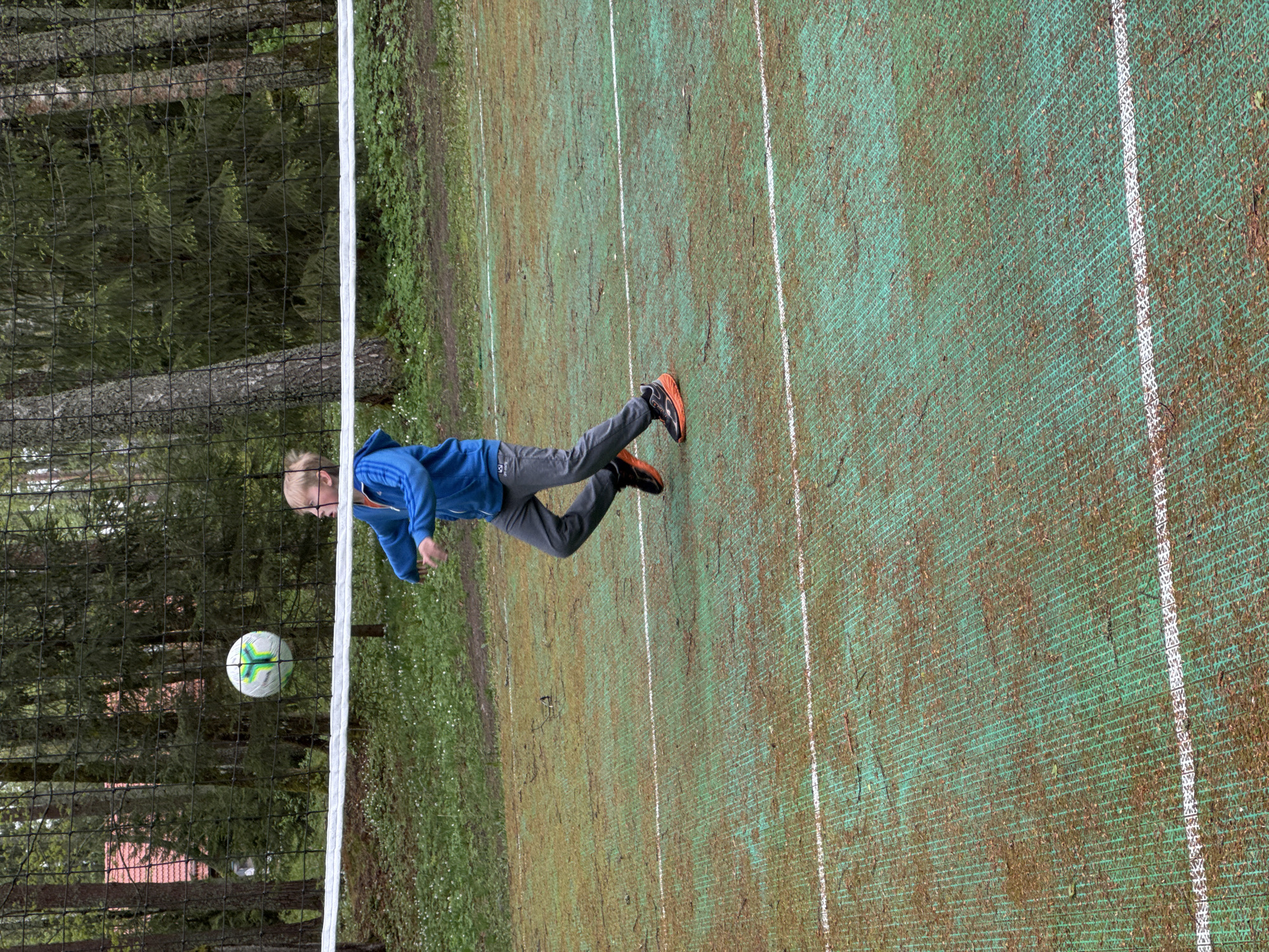 A young, light-skinned person with short blond hair wearing a blue jacket, gray pants, and orange running shoes is seen from the side, appearing to kick a soccer ball over a net. The soccer ball is white with green and black markings. The net, made of black mesh with white borders, separates a green outdoor sports field with white lines from a wooded area with trees, grass, and a building with a red roof visible through the trees. The photo appears to be taken in Aleksandrovka, Russia.