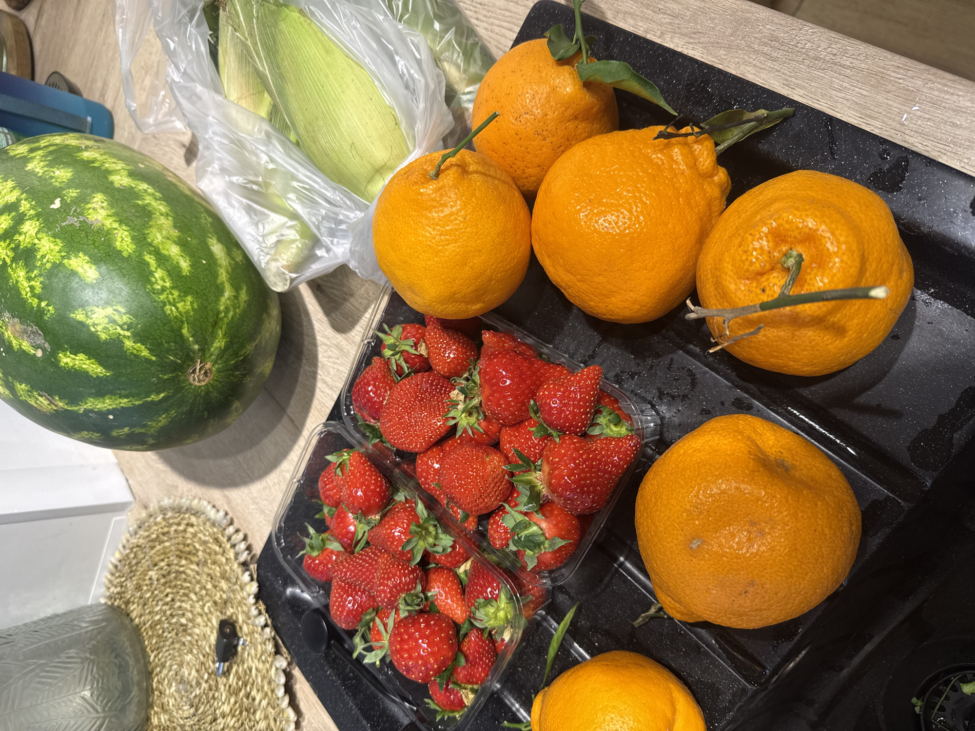 The image depicts a countertop scene with various food items. A green watermelon is situated on the left. Next to it are corn cobs in a plastic bag. To the right, there are multiple oranges, some with stems and leaves. Two plastic containers of strawberries are visible in the center. The items are arranged on a black surface that appears to be a cutting board or countertop. The scene is indoors, illuminated by ambient light. There is no evidence to suggest that this is located in Veliky Novgorod, Russia.