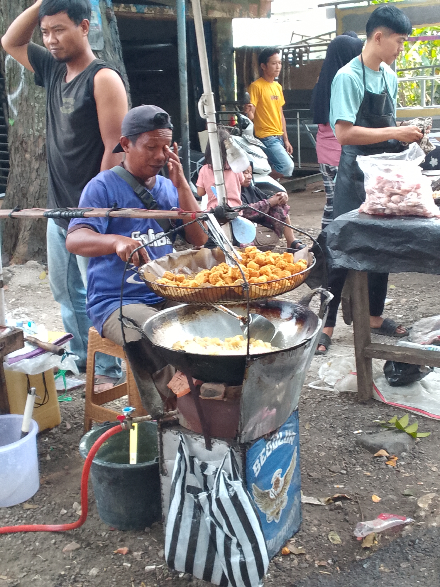 In Mandalawangi, Indonesia, a street vendor is seen preparing food outdoors. The vendor, wearing a blue shirt and a black cap, is seated on a stool and is actively frying food in a large wok. Next to him, a metal basket is filled with freshly fried items. Another man, standing behind the vendor, is wearing a black sleeveless shirt and appears to be observing. Several plastic bags, one with a distinctive eagle emblem and black and white stripes, are visible around the stall. The ground is covered with gravel and debris, suggesting a casual, possibly makeshift, market setting. A red hose connected to a valve extends from a faucet towards a black bucket. In the background, other individuals are present, including one wearing a yellow shirt and another in a blue t-shirt and apron, holding a bag of raw ingredients. The scene appears to be a typical street food vending operation.