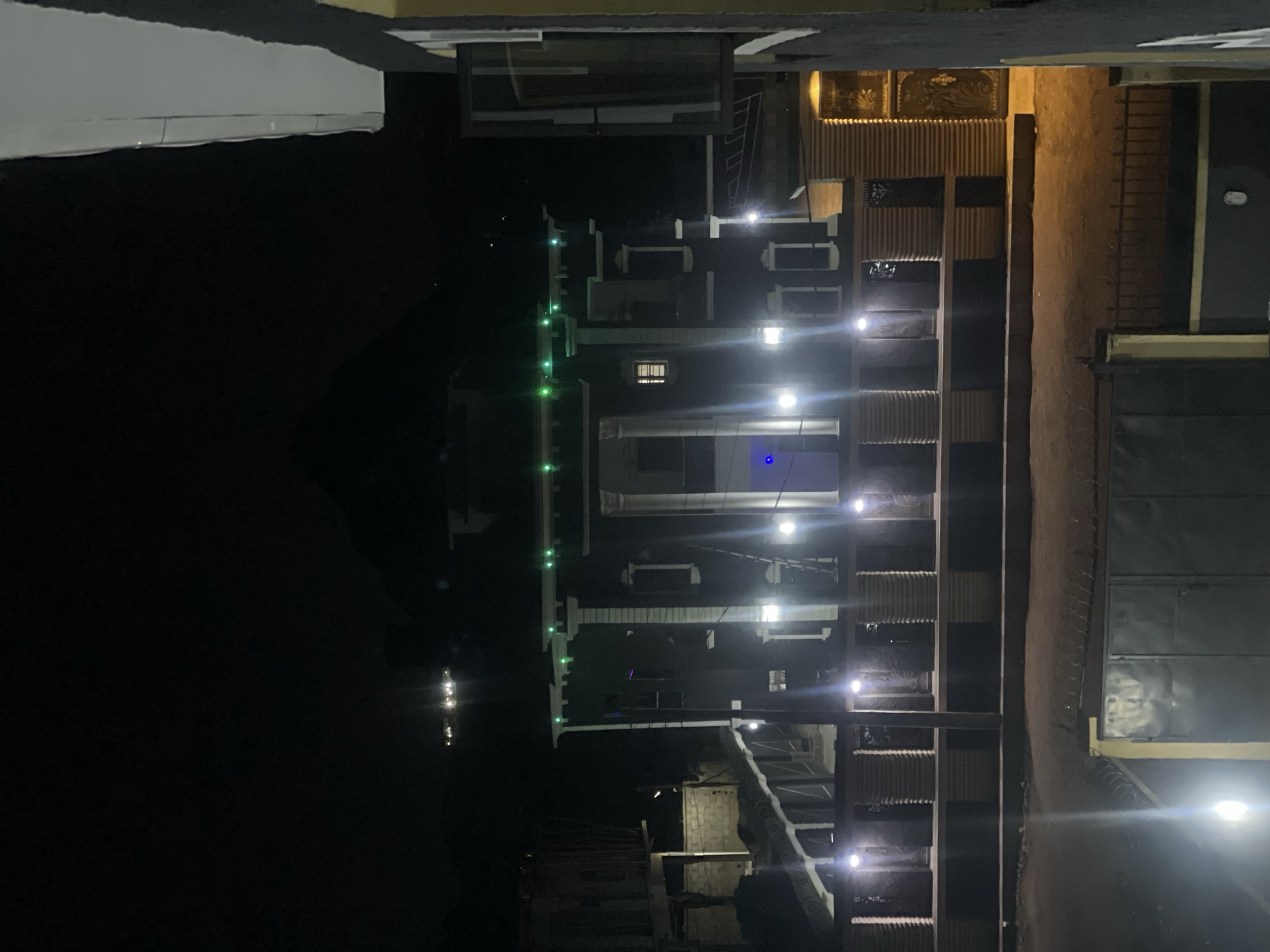 The image shows a multi-story building at night. The building has white walls, several windows, and green lights along the rooftop. There is a series of stairs with lights running up the side of the building. Adjacent to the stairs is a fence and a dirt area. Beyond the building are other structures in the dark. The location is unknown, I cannot say it is Abuja, Nigeria.