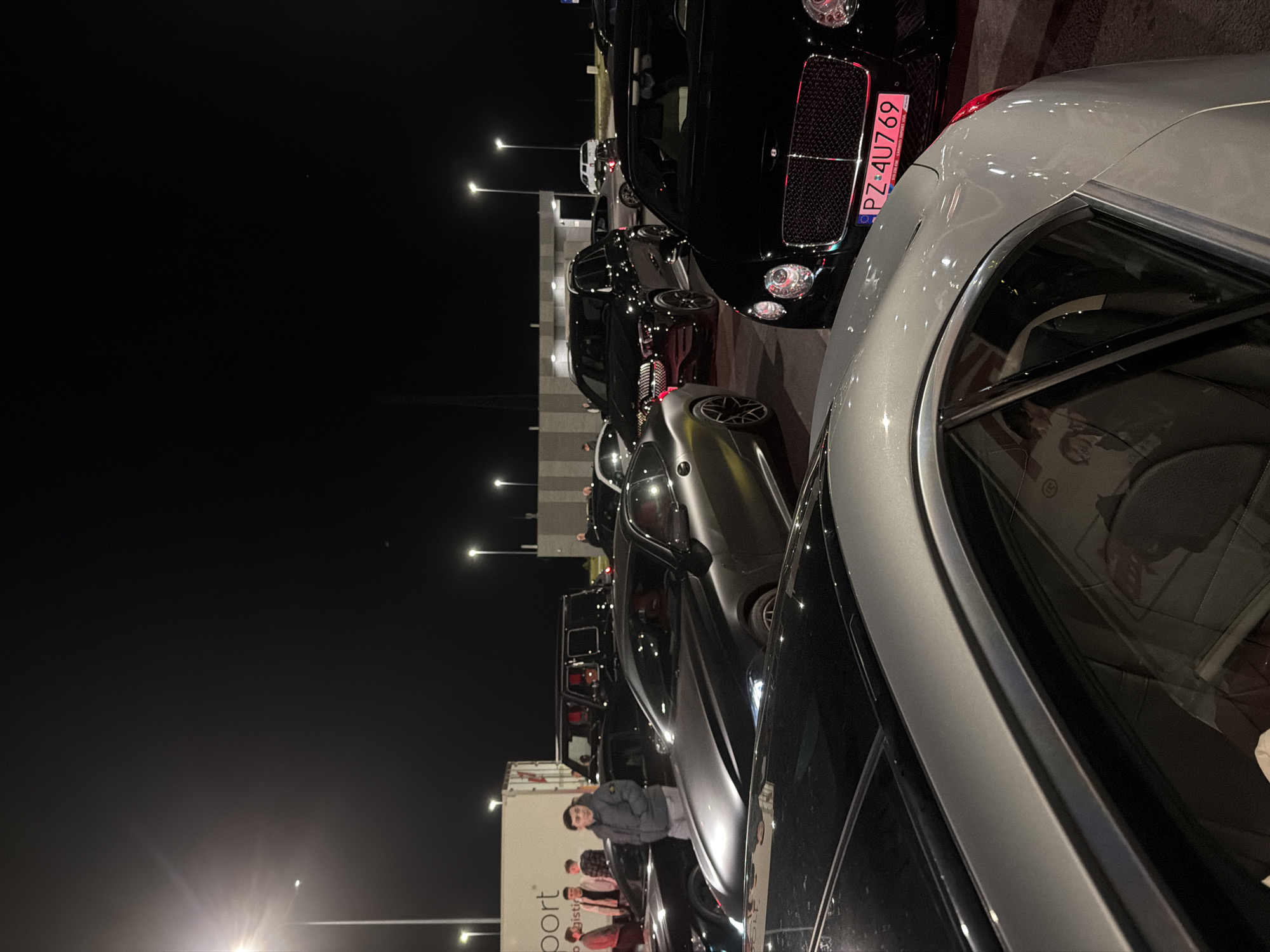 At night in Unknown, Unknown, a row of parked luxury cars, including a black Bentley with license plate PZ 4U769 and a silver car, line a road. A man in a grey jacket stands by the cars. A white truck is partially visible in the background. Streetlights illuminate the scene.