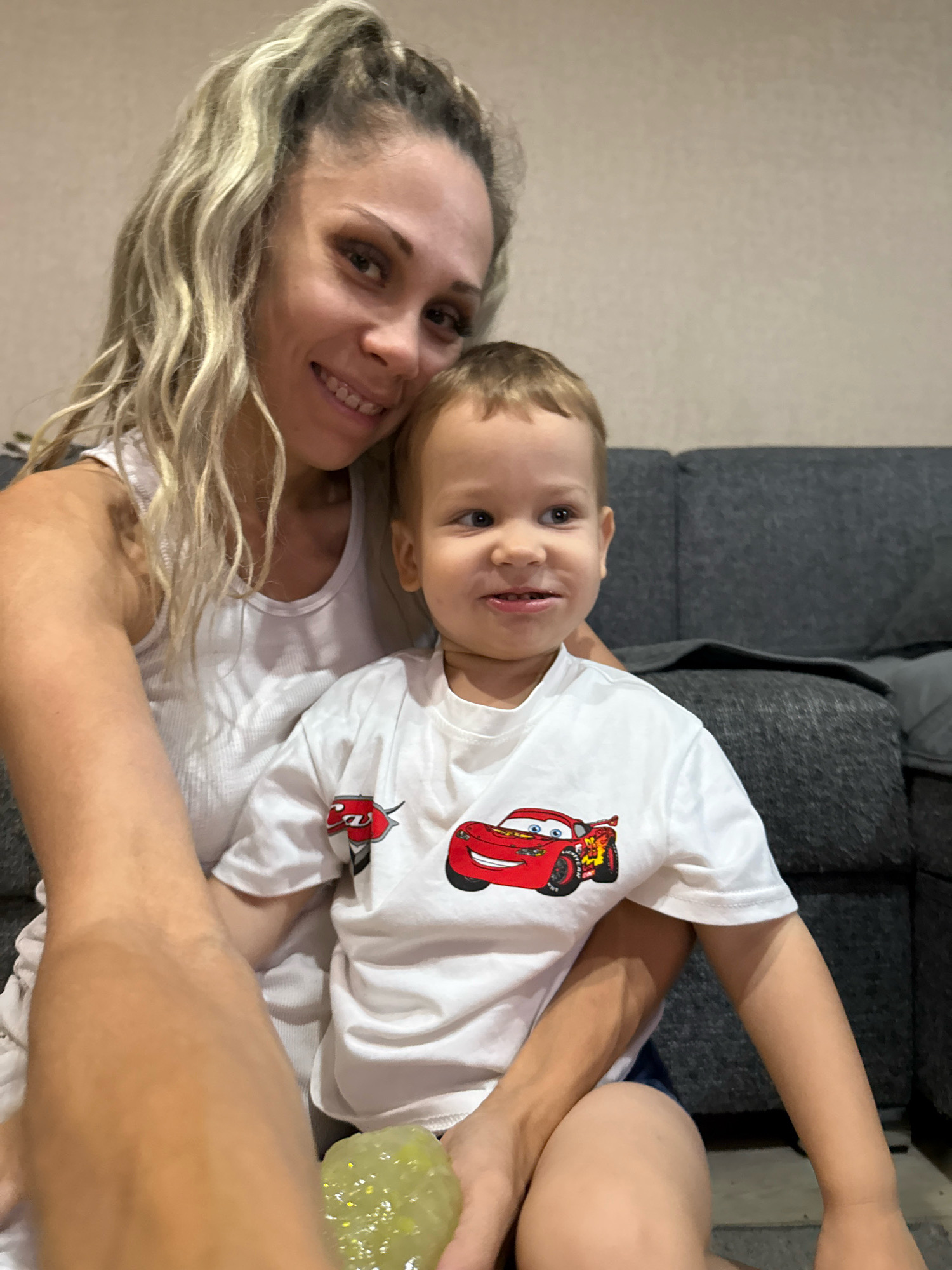 A woman with blonde hair pulled into a ponytail and a young boy with blonde hair are sitting on a couch. The woman is wearing a white tank top, and the boy is wearing a white t-shirt with a Lightning McQueen graphic. The boy is holding a green slime toy. The background appears to be an indoor setting with a textured wall. The woman is smiling and looking towards the camera, while the boy is looking off to the side. There is no discernible information to suggest the location is in Rusanovo, Russia.