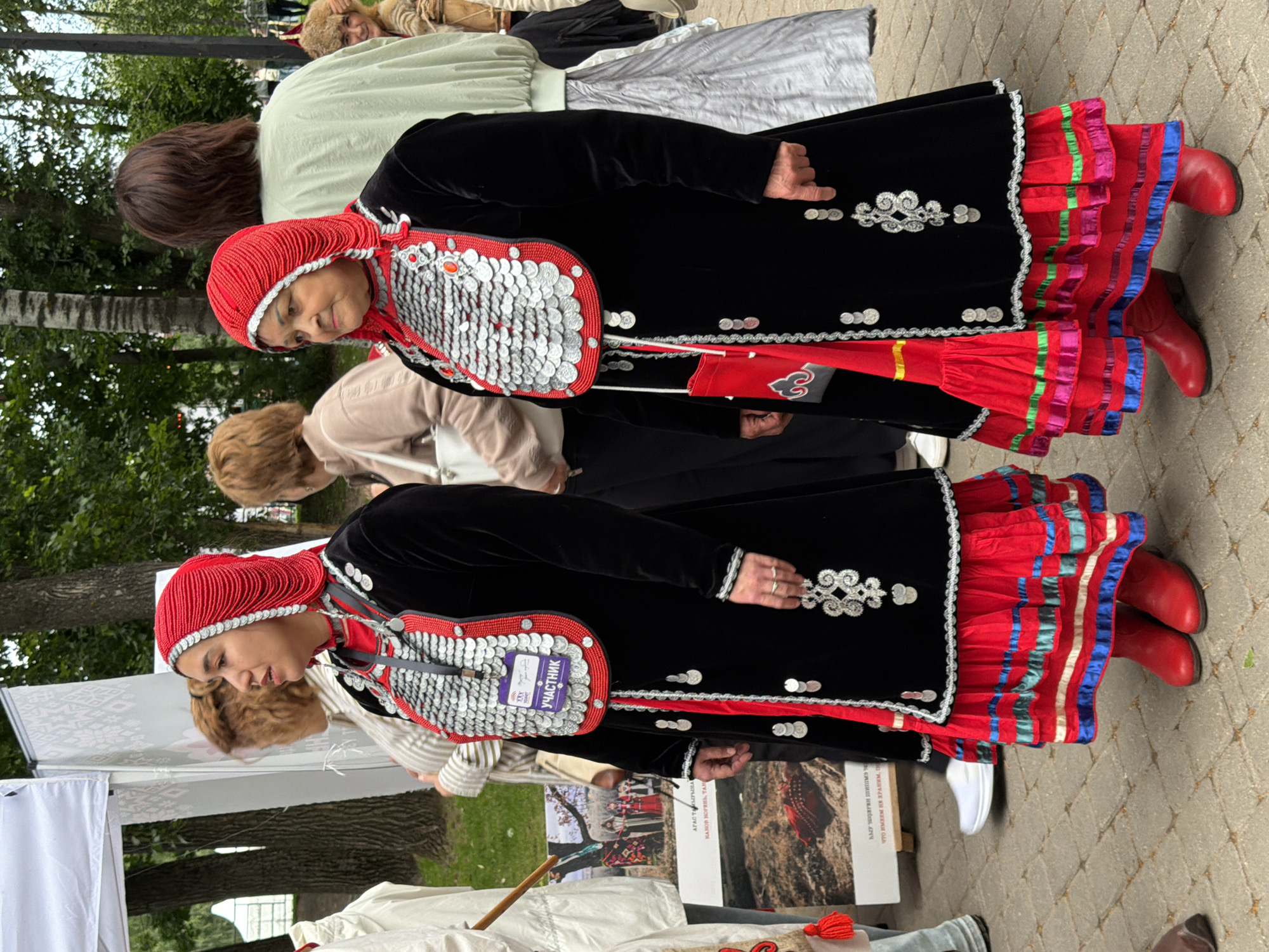 The image shows two women dressed in traditional ethnic attire, standing outdoors on a paved walkway. They are positioned in a way that suggests they are part of a cultural event or festival. The woman in the foreground, facing the left side of the frame, wears a black velvet jacket adorned with silver circular ornaments and red trim. Her head is covered with a red, beaded headdress, and she wears a red sequined chest ornament. Her lower body is covered by a vibrant red skirt with multiple ruffled layers, decorated with colorful horizontal ribbons, and she wears red boots. The second woman, behind and slightly to the right of the first, is similarly dressed, also in black velvet with silver adornments, a red headdress, and a ruffled red skirt with ribbon detailing, though her boots are not as visible.
In the background, other individuals are present, some also in traditional clothing, and trees suggest a park or outdoor gathering space. The lighting indicates it is daytime, likely a moderately overcast day given the diffused light. One woman has a purple tag with white text pinned to her chest ornament. Visible text on a poster in the lower portion of the frame appears to be in Cyrillic script. The overall atmosphere seems festive and communal, with the women showcasing their cultural heritage.