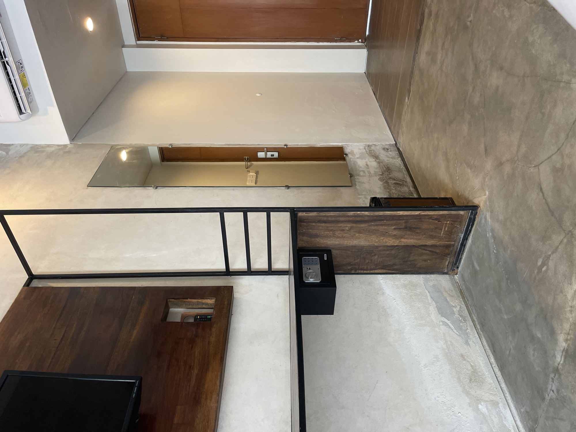 This image shows an interior space, possibly a hotel room, located in North Kuta, Indonesia. The space is defined by a concrete floor and walls with a minimalist aesthetic. The room contains a wooden structure with a black metal frame, which appears to be a bed frame or platform. A dark wood panel is attached to the frame. A black safe is positioned to the right of the bed frame. A mirror is mounted on the wall. A wooden door is located in the upper section of the image. An air conditioning unit is visible in the upper left corner. A television is partially visible on the lower left side of the image.