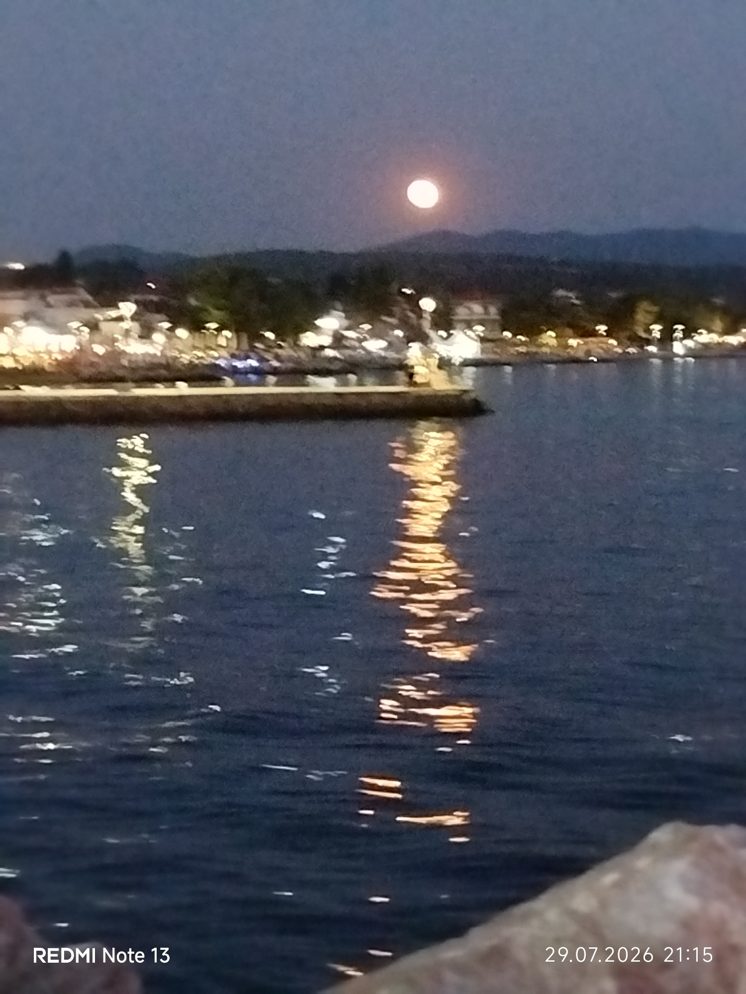 A nighttime, long-exposure photograph captures a serene view of the sea and a coastal town in Sithonia Municipal Unit, Greece. The full moon hangs low in the dusky sky, casting a soft glow. Below, the lights of the town and buildings are reflected in the calm water, creating shimmering streaks of gold and white. A dark stone pier juts out into the water from the left side of the frame. The foreground is partially obscured by a large, textured rock. The image is timestamped "29.07.2026 21:15" in the bottom right corner, and the text "REDMI Note 13" is visible in the bottom left.