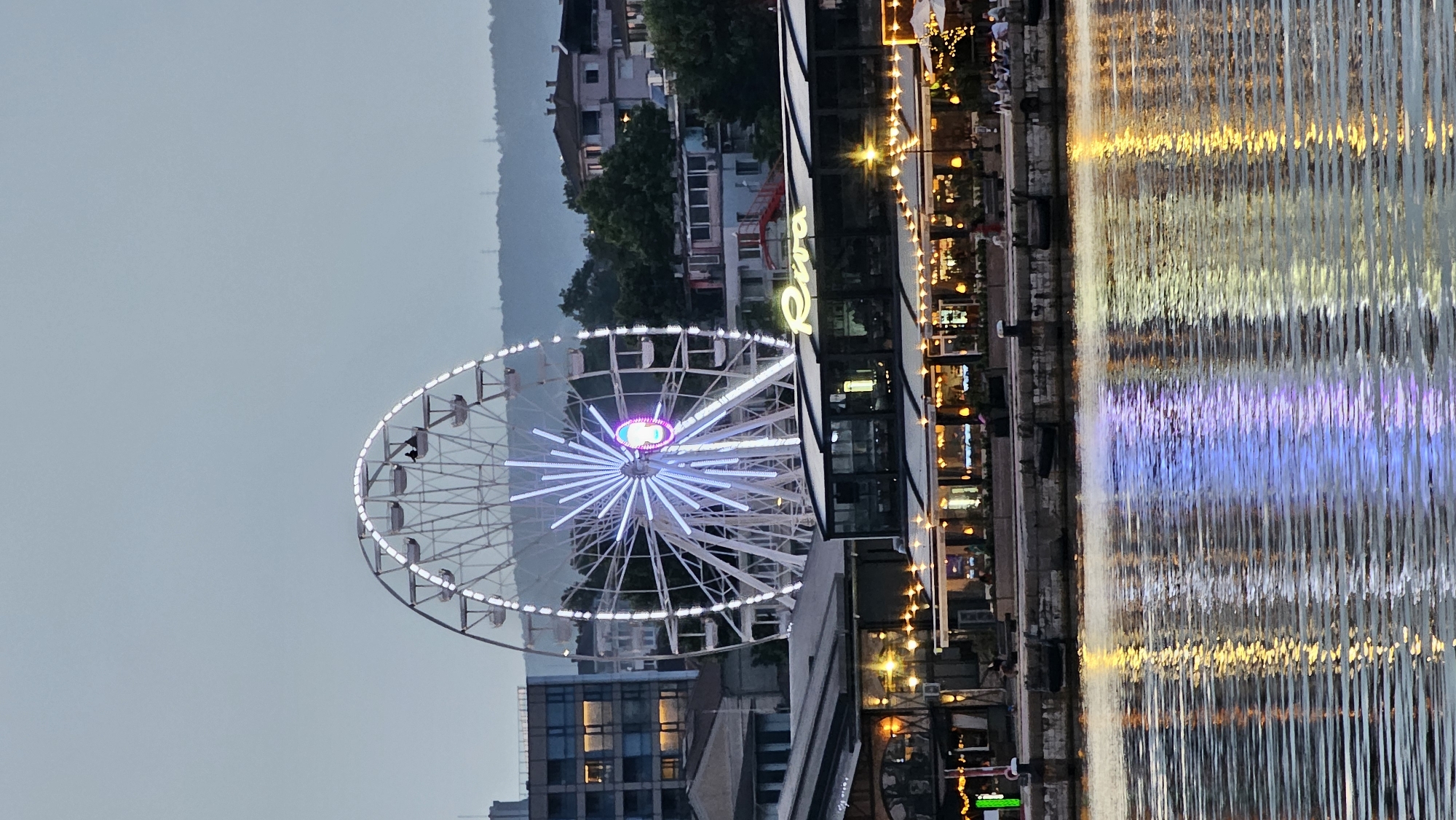 The image depicts a waterfront scene in Varna, Bulgaria, likely at dusk or early evening. Dominating the background is a large Ferris wheel illuminated with white lights, featuring a colorful, illuminated center. To the left and right of the Ferris wheel, buildings are visible, some appearing to be residential with illuminated windows, while others seem to be part of a commercial or entertainment area along the waterfront.
In the foreground, a row of establishments with covered outdoor seating areas is illuminated by numerous string lights, creating a warm, inviting ambiance. A prominent sign on one of these buildings reads "Riva" in illuminated yellow lettering. The water in the foreground reflects the lights from the Ferris wheel and the buildings, creating streaks of white, blue, and yellow on its surface.
Several individuals are visible, some seated at the outdoor tables of the waterfront establishments, others walking or standing along the waterfront promenade. Two figures can be seen in one of the Ferris wheel cabins. The overall atmosphere suggests a lively urban waterfront with recreational and dining options. The sky is overcast and a pale blue-grey, indicating the fading light of day. In the far distance, a hilly landscape with more buildings and some transmission towers is faintly visible.