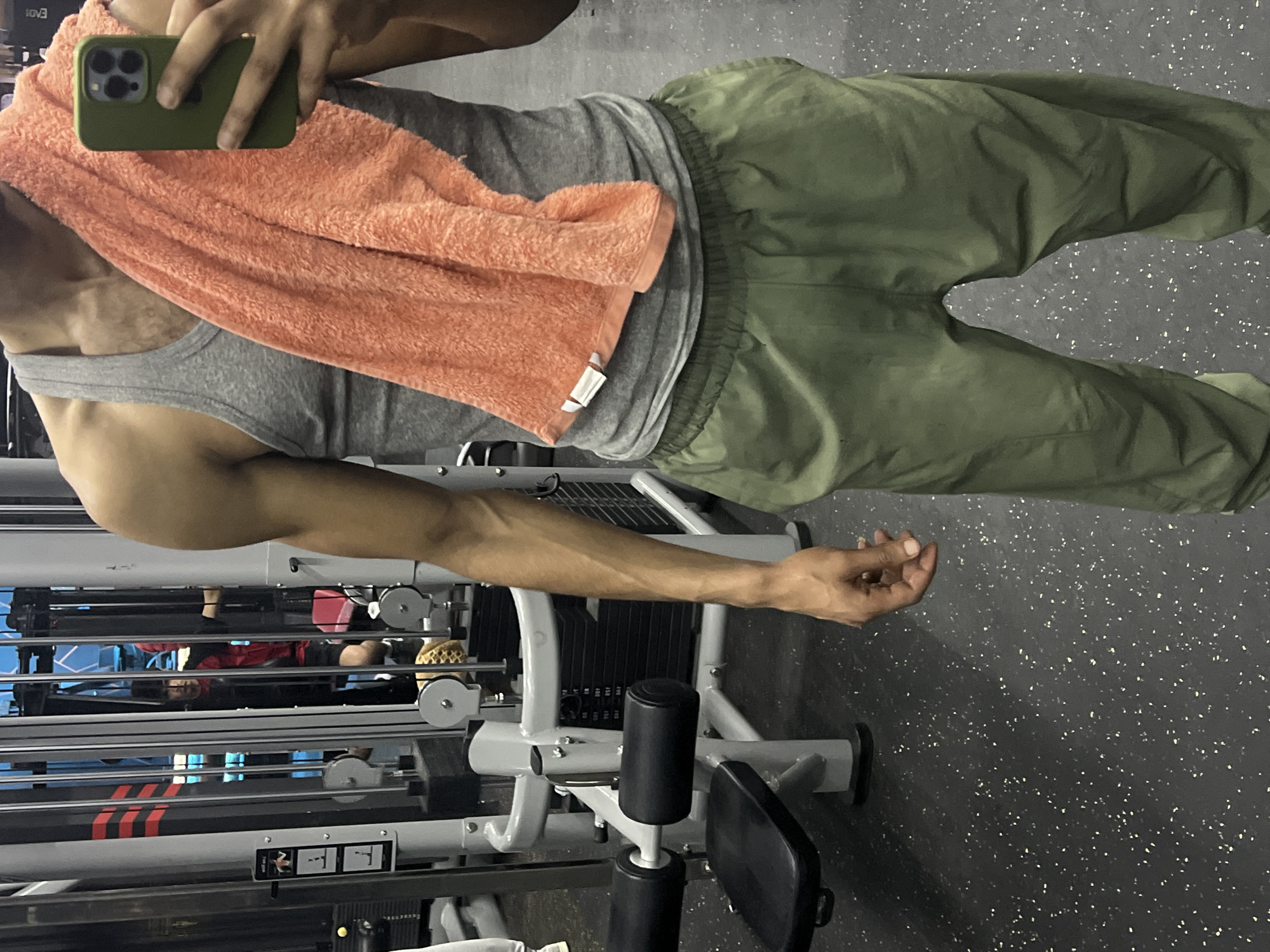 A person is taking a selfie in a gym, likely in M'Diq, Morocco. The person is wearing a gray tank top, green pants, and has an orange towel draped around their neck. They are holding an iPhone, obscuring their face. Behind them is a weight machine with black weights and a seated leg press. The floor is a speckled black and white surface. Other gym equipment and a person wearing red are visible in the background, reflected in the machine's frame.