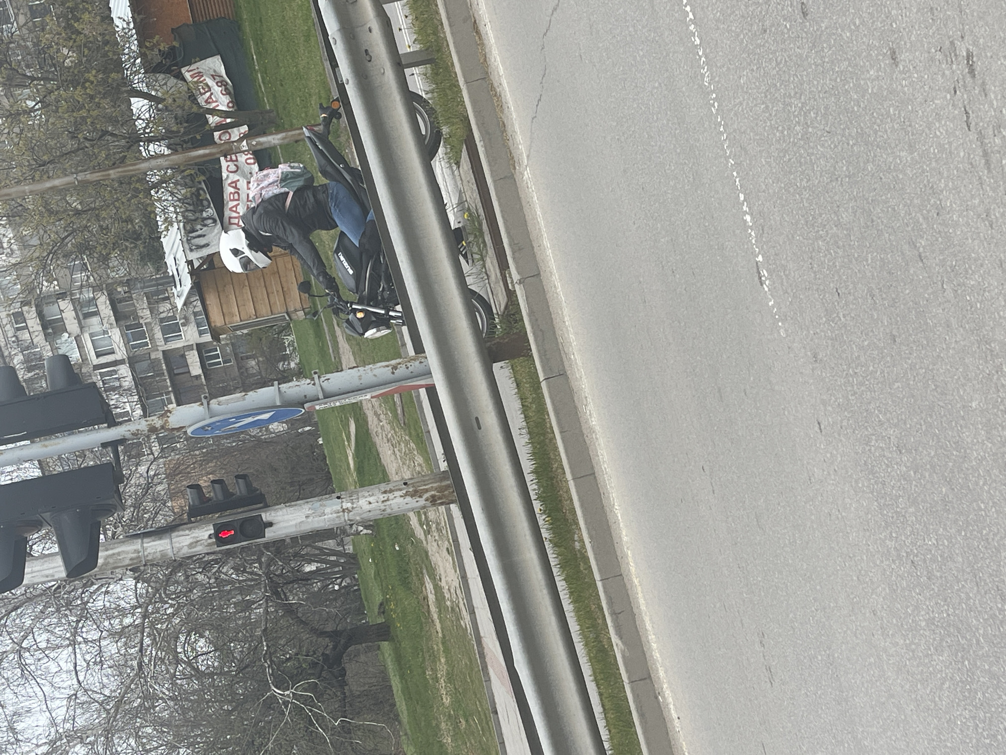The image shows a person wearing a helmet and riding a Suzuki motorcycle on a street with a sidewalk in Sofia, Bulgaria. The person is wearing a dark jacket and blue jeans. The road is asphalt, and there is a metal guardrail separating the road from the sidewalk. The traffic light shows a red light. A sign is attached to a building in the background. The location appears to be near an apartment building.