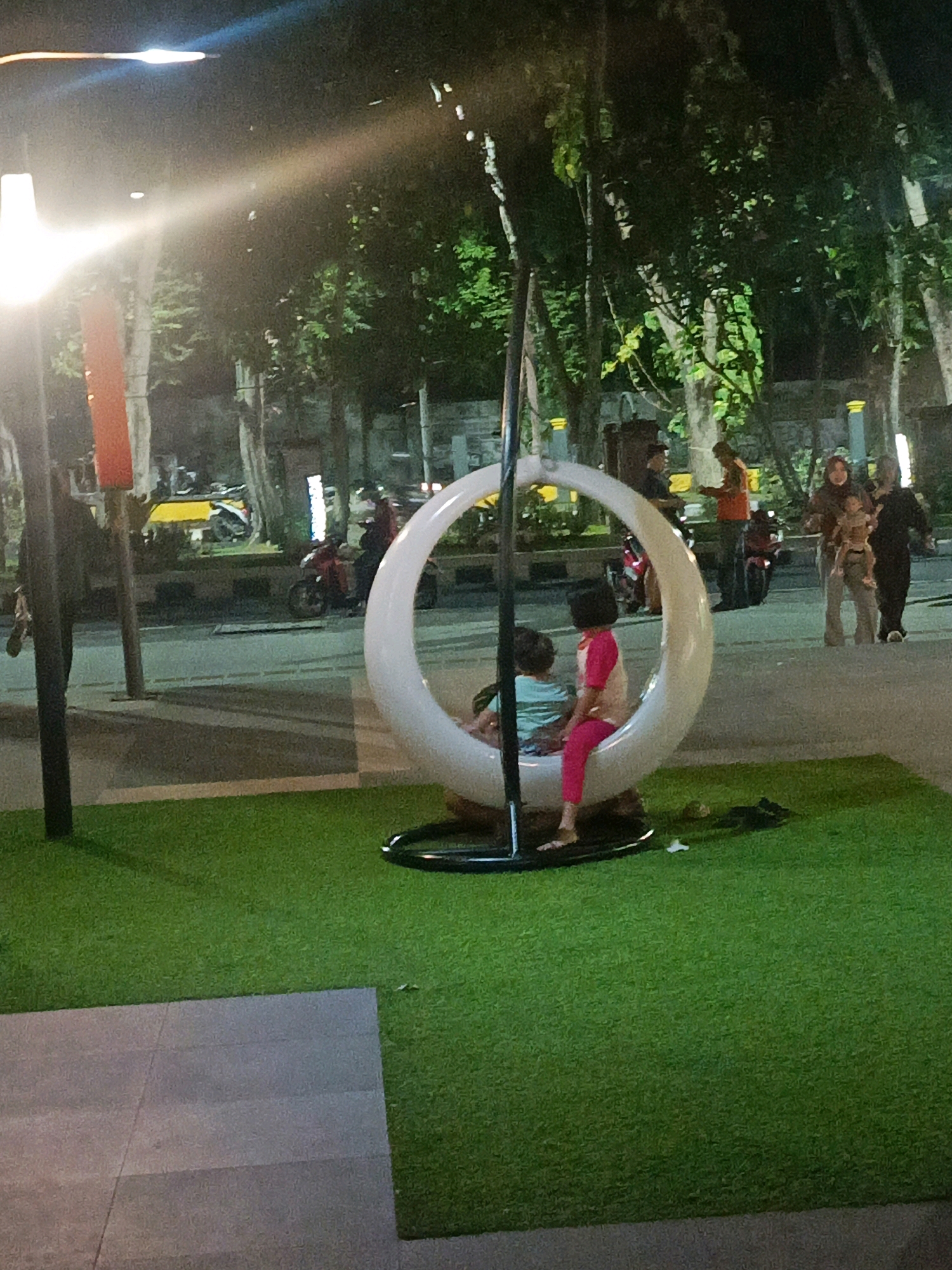 The image shows a nighttime scene in a park in Sidoarjo, Indonesia. In the foreground, a white, crescent-shaped swing is occupied by two young children. One child, wearing pink pants and a pink and white shirt, sits facing forward, while another child, wearing a light blue shirt, is seated behind. The swing is situated on an artificial green turf area. Behind the swing, a paved walkway is visible, with trees and streetlights lining the background. Several people are seen in the mid-ground and background, including adults with children, and some on motorcycles. The atmosphere appears to be relaxed and social, suggesting a public gathering space. The lighting is dim, consistent with a nighttime setting, with artificial lights illuminating the park area.