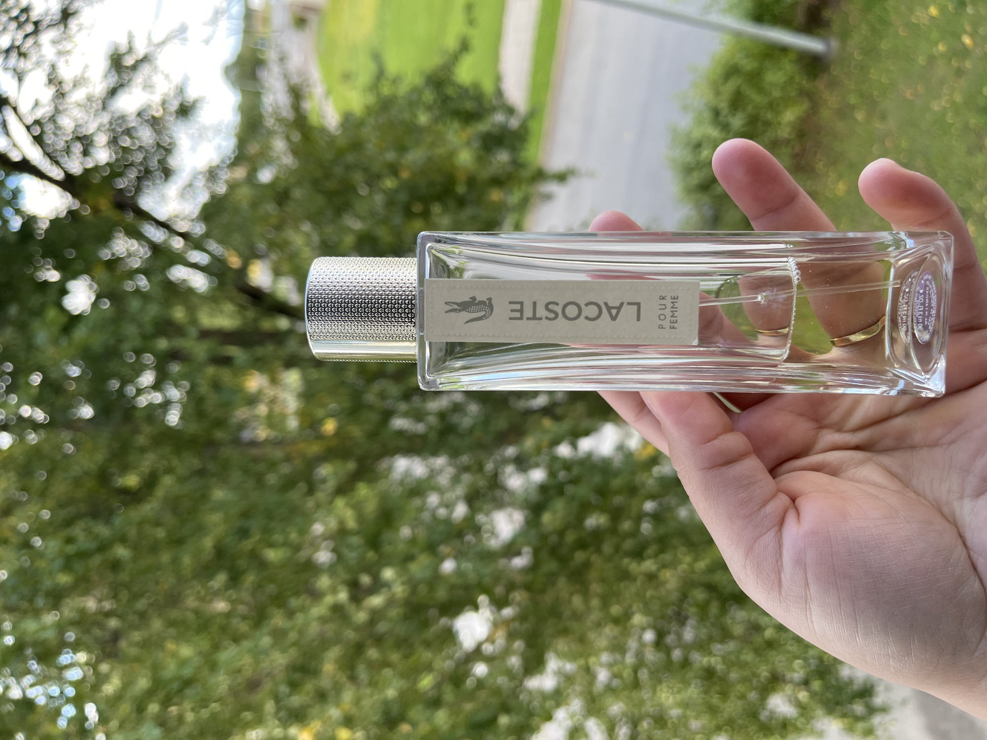 A hand holds a clear glass bottle of Lacoste Pour Femme perfume in front of a blurred background of green trees and a grassy area. The bottle has a silver textured cap and a light-colored label with the Lacoste logo and text. The background suggests an outdoor setting, possibly a park or residential area with foliage and a road visible. The lighting is natural and appears to be daylight. The image is focused on the perfume bottle and the hand holding it.