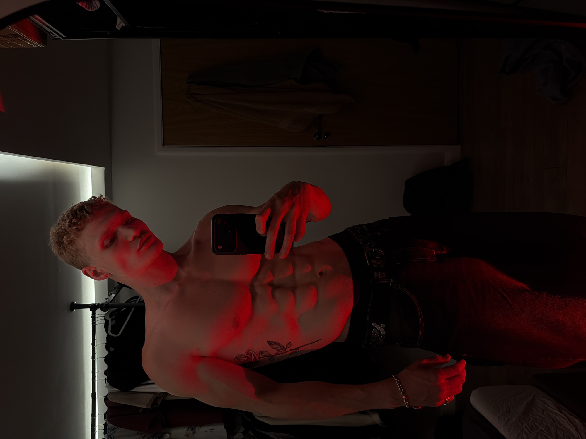 A shirtless young Caucasian male with short blonde hair stands in a dimly lit room, posing for a mirror selfie. He is illuminated by a vibrant red light, which casts a strong glow on his muscular physique, particularly his abdominal muscles and a rose tattoo on his left side. He is wearing dark denim jeans with a studded belt. The background shows a closet with clothes hanging on a rack and a wooden door with a towel draped over it. The floor appears to be wood. The scene is set in a residential room, with no specific indicators of its location within London, United Kingdom.