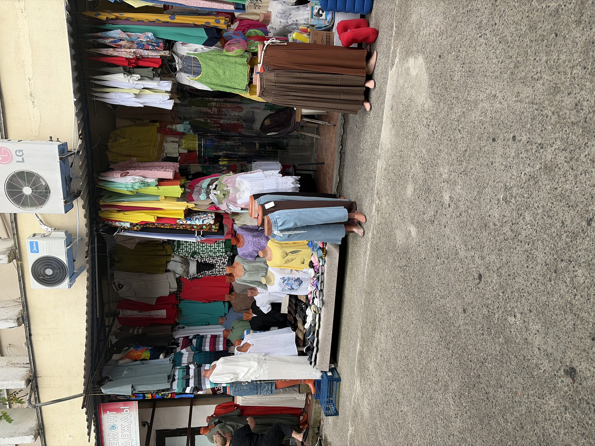 This is a photograph of an outdoor clothing stall in Sochi, Russia, located on a street with a concrete surface. The stall is covered by a corrugated metal awning, and it displays a variety of women's clothing, including dresses, skirts, and tops. The clothing is hung on racks and displayed on mannequins. There are also shoes on display. In the background, there is a building with an air conditioning unit mounted on it. A woman is visible on the left side of the frame, reaching towards a rack of clothing. The scene is brightly lit, suggesting it is daytime.