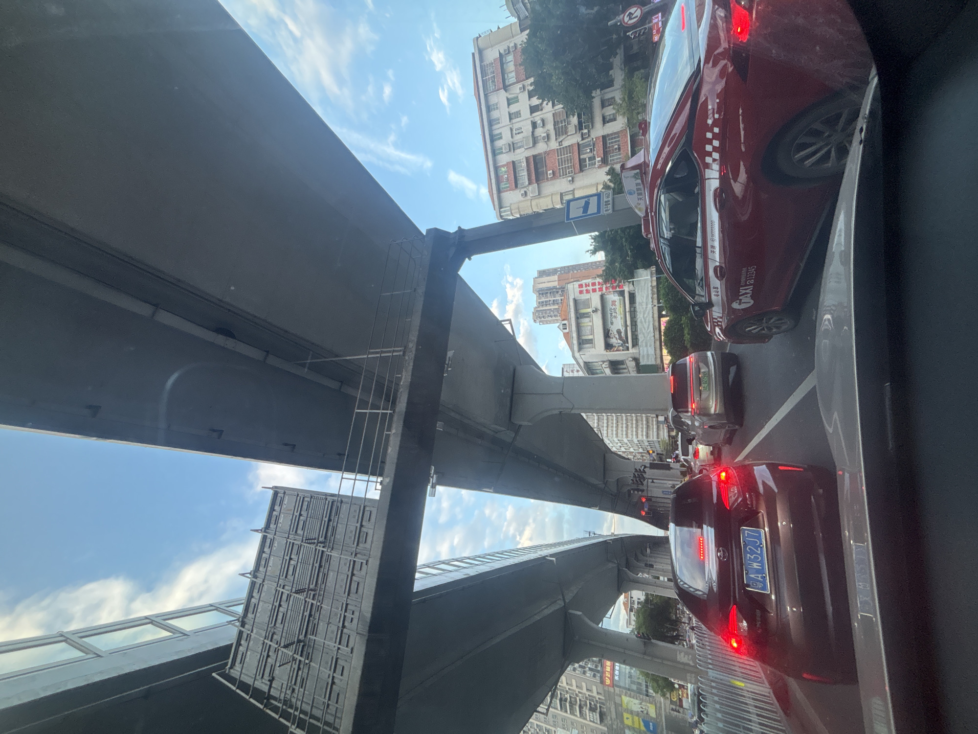 The image depicts a street-level view from inside a vehicle, looking forward through the windshield and out at a busy urban traffic scene. The foreground shows the dashboard and hood of the vehicle the photographer is in. Directly ahead is a dark-colored sedan with a license plate reading "A W32J7". To the right of this vehicle is a red taxi with "TAXI" branding and Chinese characters on its side. The taxi also has a rooftop sign. The road ahead is congested with other vehicles, including a silver SUV. Overhead, a massive elevated structure, likely an overpass or elevated train line, dominates the sky, supported by large concrete pillars. The sky is blue with scattered white clouds. In the background, multi-story buildings line the street, some appearing residential with visible air conditioning units. Traffic lights are visible, indicating an intersection. The overall impression is one of a densely populated urban environment in China, possibly the Liwan District, with significant infrastructure supporting transportation.