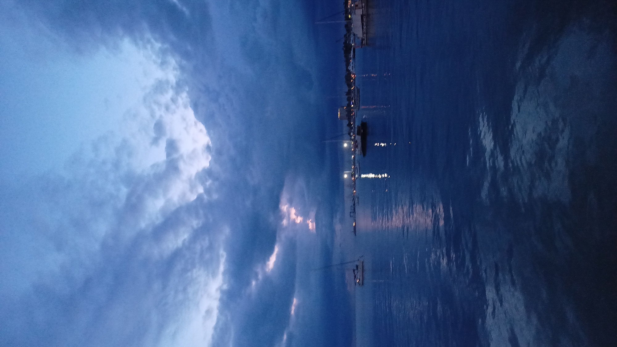 The image captures a dusk or pre-dawn scene over the Adriatic Sea in Umag, Croatia, likely within its harbor or marina. The upper portion of the frame is dominated by heavy, dark blue-grey clouds. A narrow, irregular vertical opening in the central cloud mass reveals streaks of faint pink and purple light, suggesting light from a distant sun breaking through the overcast sky. The lower half of the image consists of calm, dark blue water, reflecting the sky and distant light sources. Numerous boats are moored across the water; a light-colored sailboat or catamaran is visible on the far left, and a dark-hulled boat with a mast is positioned towards the center-right. In the mid-ground, the distant shoreline of Umag is delineated by an array of small, bright orange and white city lights, which cast elongated reflections upon the water's surface. The water exhibits subtle ripples. No individuals are visible in the scene.
