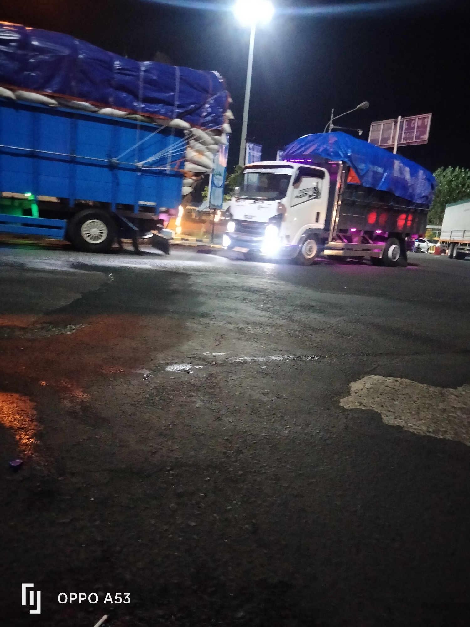 In Melaya, Indonesia, at night, two trucks are parked on a wet, dark asphalt surface. The truck on the left is blue with a blue tarp covering its cargo, which appears to be sacks of grain. It has a white rim on its front wheel and green undercarriage lights. The truck on the right is white with a black flatbed and a blue tarp. It has purple accent lights along the bottom of its bed and red taillights. Its headlights are illuminated. In the background, a street light shines brightly, and traffic signs are visible. Trees are also present in the background, and the scene is illuminated by artificial lights. The overall impression is of a busy industrial or transportation hub at night.