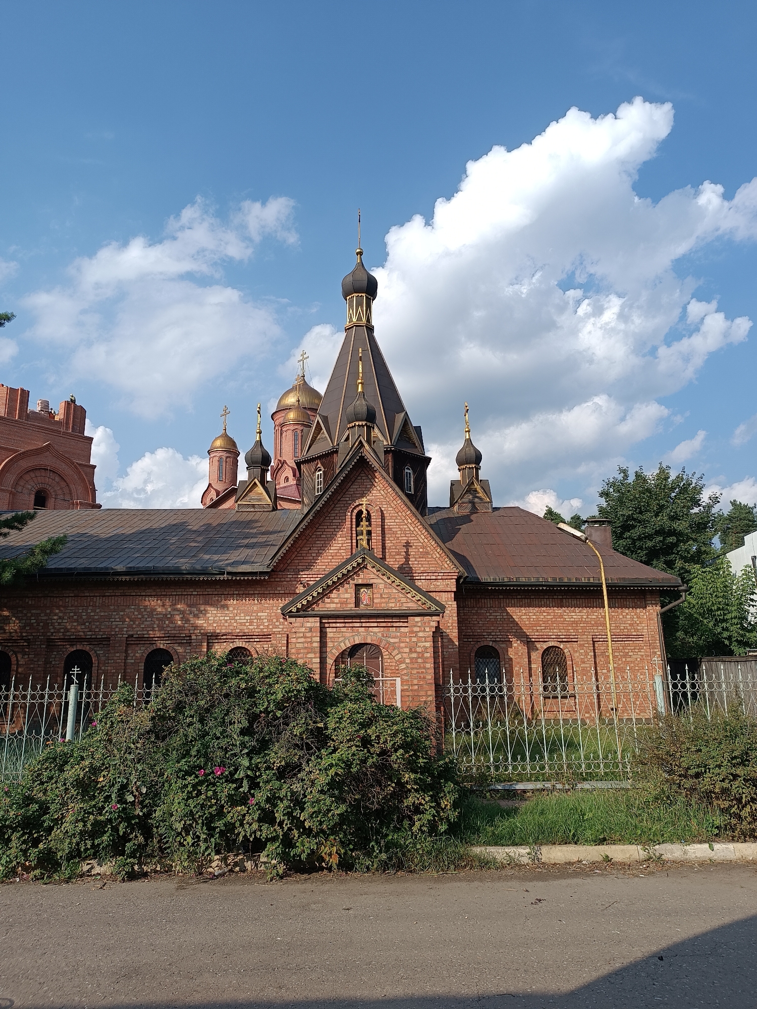The image depicts the Church of the Intercession of the Holy Virgin, located at 15 Tsentralnaya Street in Il'inskiy, Russia. The red brick church features multiple domes, including a central large dome and several smaller domes. The building exhibits traditional Russian Orthodox architecture with arched windows and decorative brickwork. In the foreground, a dense green bush with some pink flowers is visible, separated from the church by a white decorative fence. Beyond the fence, there is a strip of green grass and a paved road. The sky above is blue with scattered white clouds. Trees are visible to the right of the church. No people are present in the image.