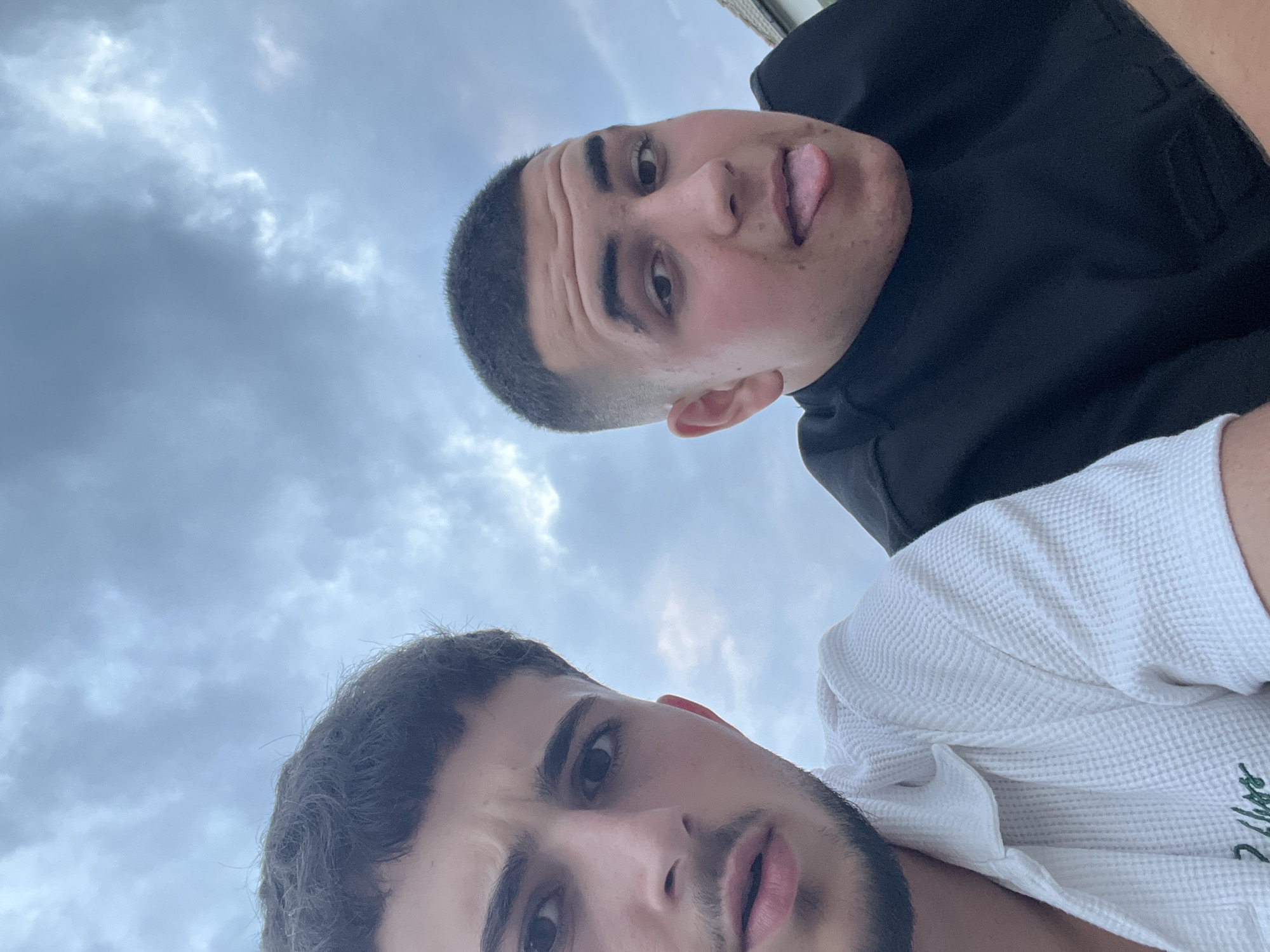 The image is a selfie taken outdoors with a cloudy sky in the background. Two young men are visible, both appearing to be in their late teens or early twenties. The man on the left has dark hair, a short beard, and is wearing a white waffle-knit shirt with a green embroidered logo. He is looking directly at the camera with a slightly furrowed brow. The man on the right has a buzz cut and is sticking his tongue out at the camera. He is wearing a black t-shirt. The perspective suggests they are sitting or leaning against something, possibly outdoors in a residential area. No specific urban features of Barutin, Bulgaria, are identifiable in the provided image or crops.