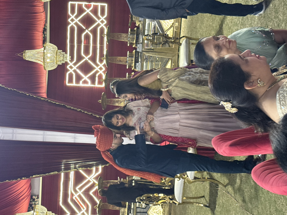 A group of people, seemingly at an event like a wedding, are standing and talking. A man in a turban is looking at a phone held by a woman. There are also two women looking off to the right. The background consists of red drapes, chandeliers, and dining tables with gold serving dishes. The scene is warmly lit, suggesting an evening gathering.