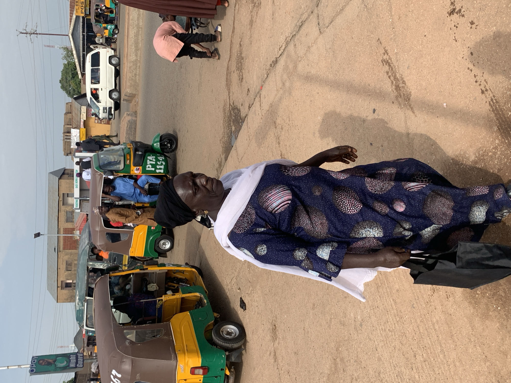 The image captures a street scene in Gura Top, Nigeria, focusing on a woman walking towards the viewer. She is wearing a dark blue dress with a white scarf draped over her shoulders and head, and she carries a black shopping bag in her right hand. Her expression is neutral as she looks slightly to her left. Behind her, a busy intersection is visible with several auto-rickshaws, locally known as "Keke NAPEPs," and a white van. One Keke NAPEP, a green and yellow model, is parked in the foreground to the left. Another auto-rickshaw, also green and yellow, is further back, as is a white van. People are seen in and around these vehicles, suggesting a transportation hub. Buildings line the background, some with signs indicating shops or services. The ground is a dusty, reddish-brown surface with some scattered debris. The overall impression is one of everyday life and transportation activity in a Nigerian city.