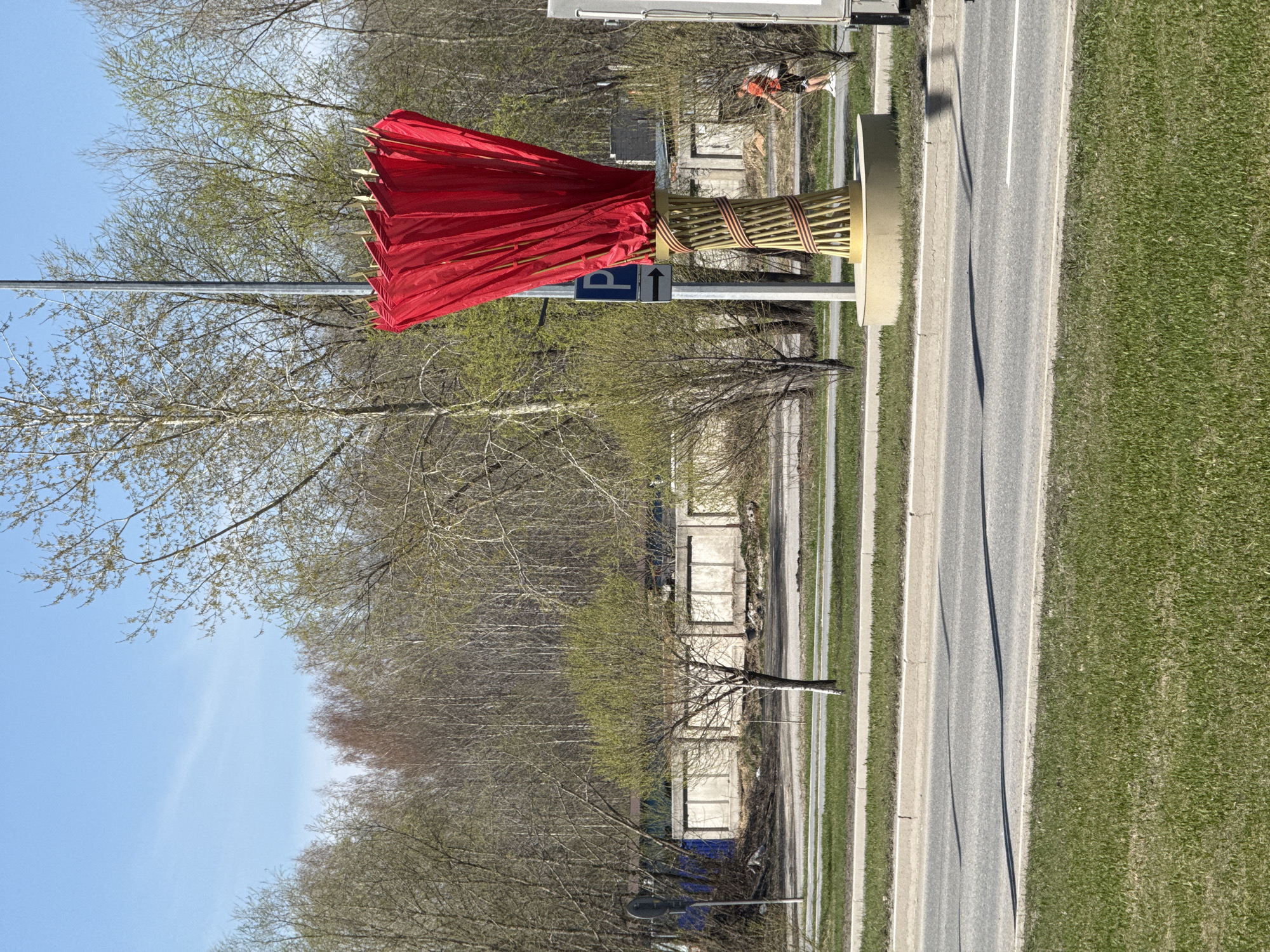 A roadside installation featuring a red cloth attached to a bundled stick structure is shown on a road in Tobolsk, Russia. The installation is adjacent to a road with a grassy shoulder. Bare trees and a blue sky are visible in the background. A person is working in the far background. A parking sign is affixed to the supporting post of the art installation.