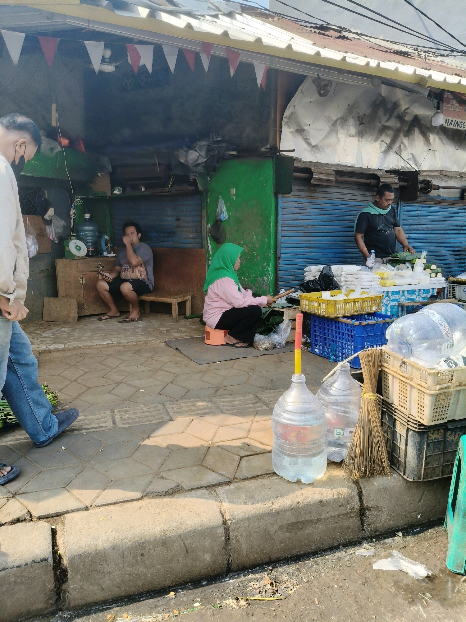 The image depicts an outdoor market scene in South Tangerang, Indonesia. A man wearing a black t-shirt and a light green scarf is standing behind a table laden with food items, likely being prepared for sale. He holds a bottle of water and appears to be interacting with customers or arranging goods. To his left, a woman in a pink shirt and green hijab is seated on an orange stool, also engaged with food preparation, using a wooden utensil to wrap items in leaves. In the foreground, several large water bottles and a broom are placed on the ground. The market is situated along a sidewalk with a concrete curb, and various crates and baskets are visible, containing more food products and supplies. A man in a white shirt and jeans is walking away from the camera on the left side of the image. In the background, a simple structure with a corrugated metal roof and blue shutters suggests the market is a semi-permanent setup. The overall environment appears to be a bustling street market in an urban setting.