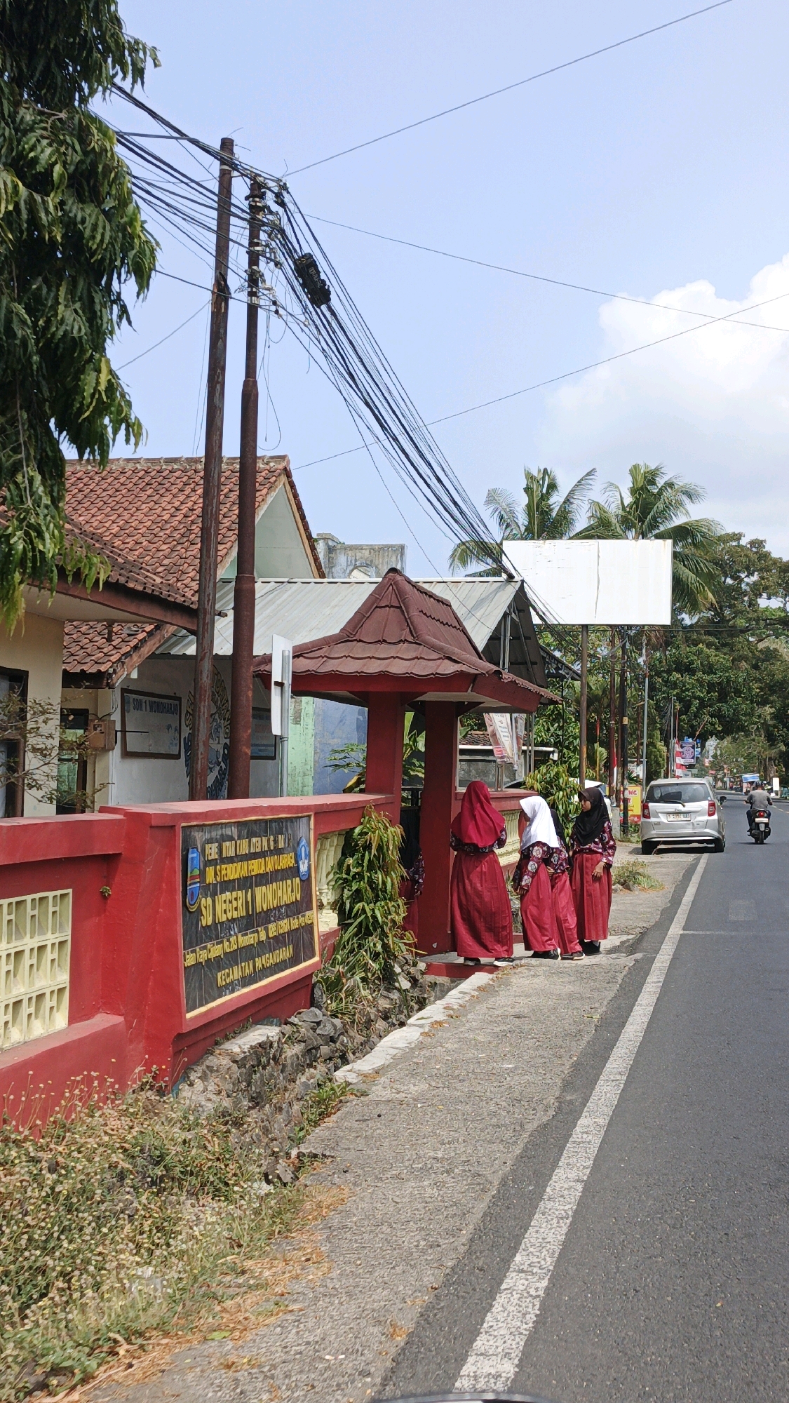 The image shows the front entrance of SD Negeri 1 Wonoharjo, a primary school located in Desa Wonoharjo, Indonesia. A red gate and fence line the property, with a red-roofed entrance shelter. Three young girls, dressed in red school uniforms with headscarves, stand on the sidewalk near the entrance. They appear to be waiting or observing something. A silver car is parked on the side of the road, and a motorcycle is visible further down the street. The environment is semi-urban, with trees and buildings visible in the background. Power lines are strung across the sky, supported by utility poles. The road is paved with asphalt and has a white line marking the lane. The sky is clear with some scattered clouds.