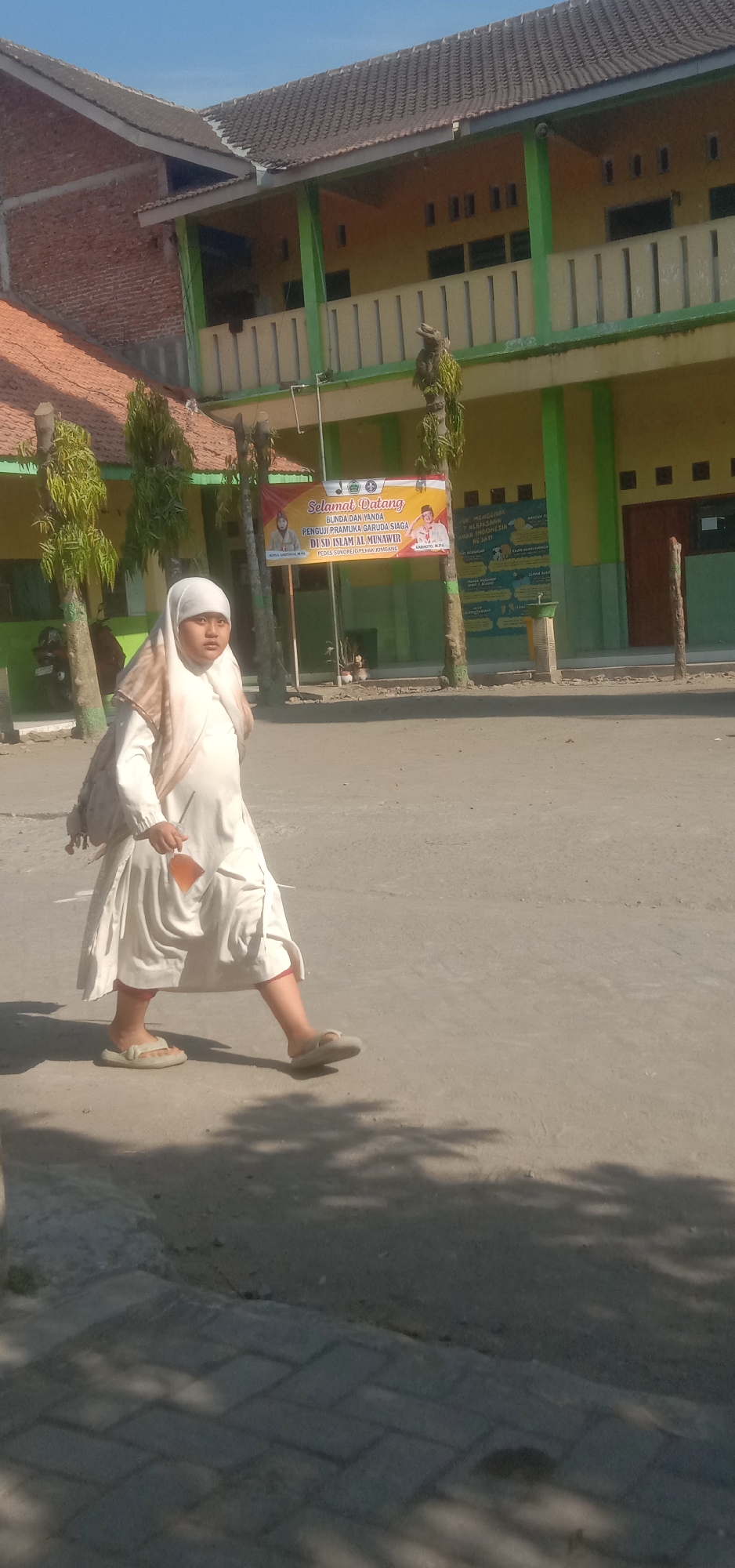 In Sukorejo, Indonesia, a young girl wearing a white hijab and a white dress walks along a paved path in front of a school building under a clear blue sky. The school building appears to be a two-story structure with a tiled roof and green accents on the exterior. A banner is visible on the building, which appears to be a welcome message for a school event. The girl is carrying a backpack over her shoulder and appears to be holding a small orange object in her right hand. The ground is a mix of asphalt and paving stones, with shadows from trees cast across the scene. The overall impression is that of a typical day at an Indonesian school.