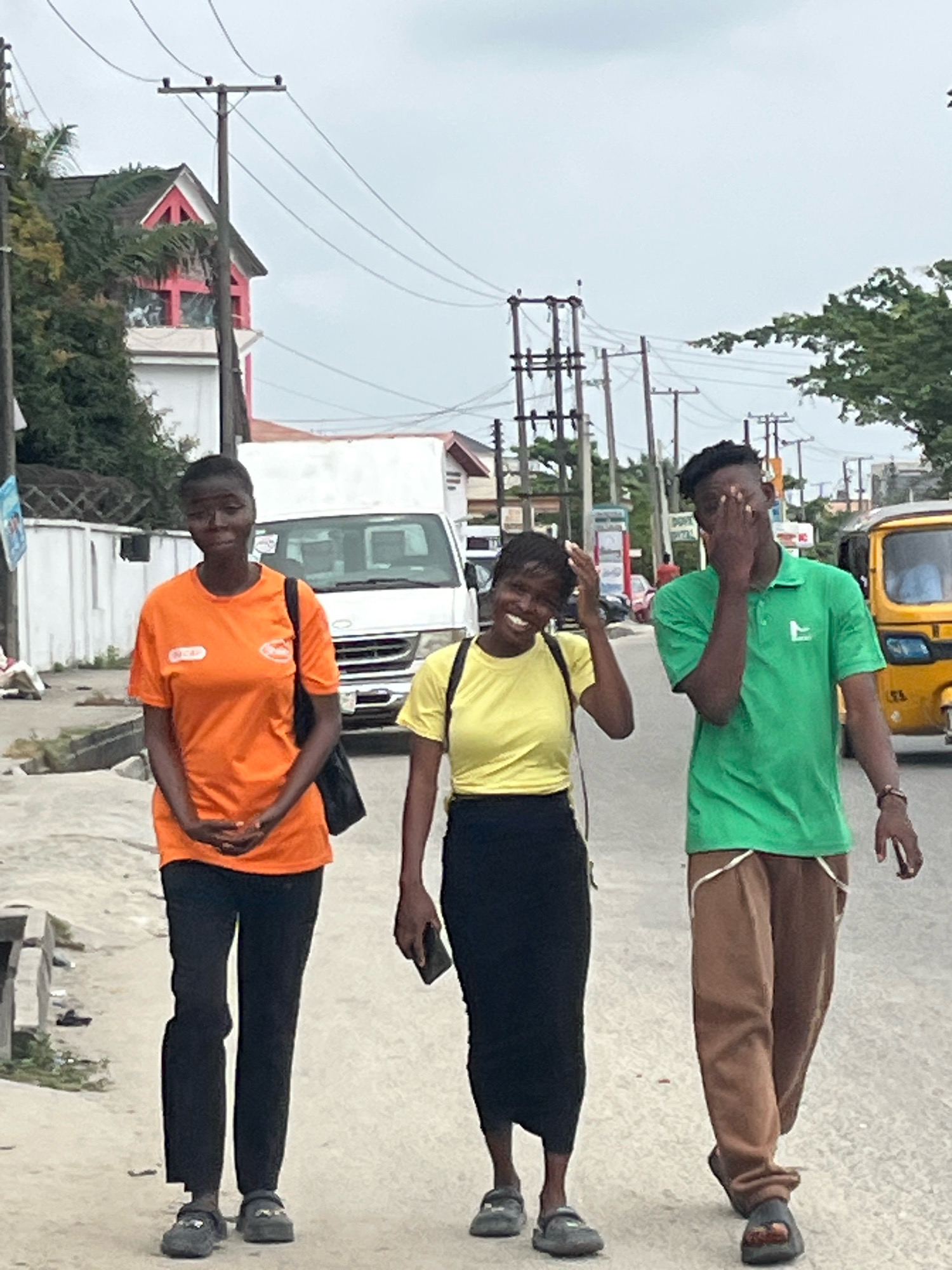Three young adults are walking on a paved street in Ado, Nigeria. The person on the left, a woman, is wearing an orange t-shirt and black pants, carrying a black bag over her shoulder. In the middle, a woman in a yellow t-shirt and black skirt is smiling and touching her head. To the right, a man in a green t-shirt and brown pants is covering his face with his hand and looking away. In the background, a white van, a yellow auto-rickshaw, and several utility poles with overhead wires are visible. Trees and buildings line the street. The sky is overcast.