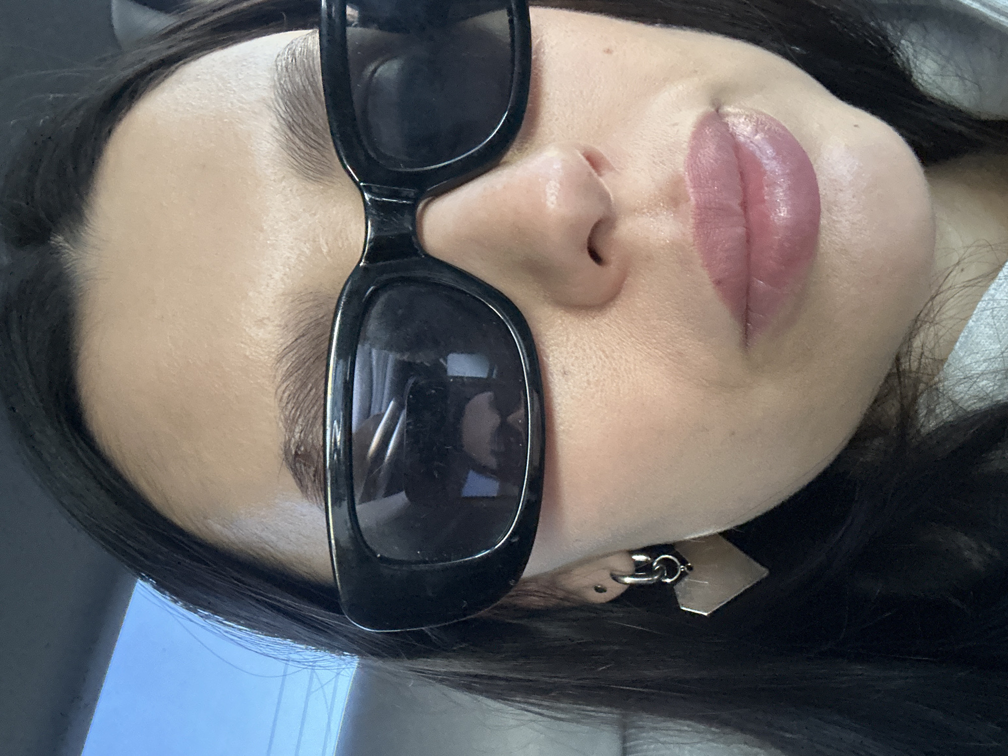 A close-up selfie of a woman's face tilted to the left, featuring black sunglasses with rectangular frames. The focus is on the upper portion of her face, showcasing a fair complexion, dark hair, full pink lips, and a nose. Part of an earring is visible. The reflection in the sunglasses lenses shows a smartphone and possibly the interior of a vehicle. The location is unspecified and no information is available regarding the city of Chaikovsky, Russia.