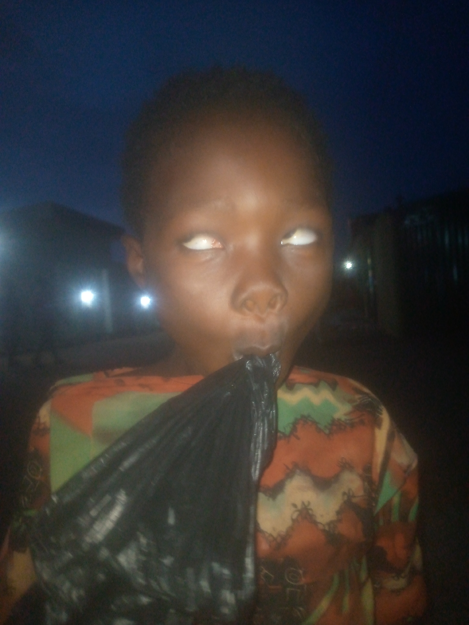 A close-up, dimly lit photo shows a young child, likely a boy, from the chest up, engaged in a playful interaction with a black plastic bag at night in Ikorodu, Nigeria. The child has dark skin and short, dark hair. He is wearing a colorful patterned shirt with shades of orange, green, and brown/black. His eyes are rolled upwards, revealing only the whites, which gives his expression a dramatic or humorous quality. A crumpled black plastic bag is positioned near or in his mouth, suggesting he might be blowing into it or playfully biting it. The background is very dark, indicating nighttime, with several bright, blurred artificial lights visible in the distance, likely from buildings or streetlights. The overall scene appears spontaneous and lighthearted, capturing a moment of childish play. No text is visible in the image.