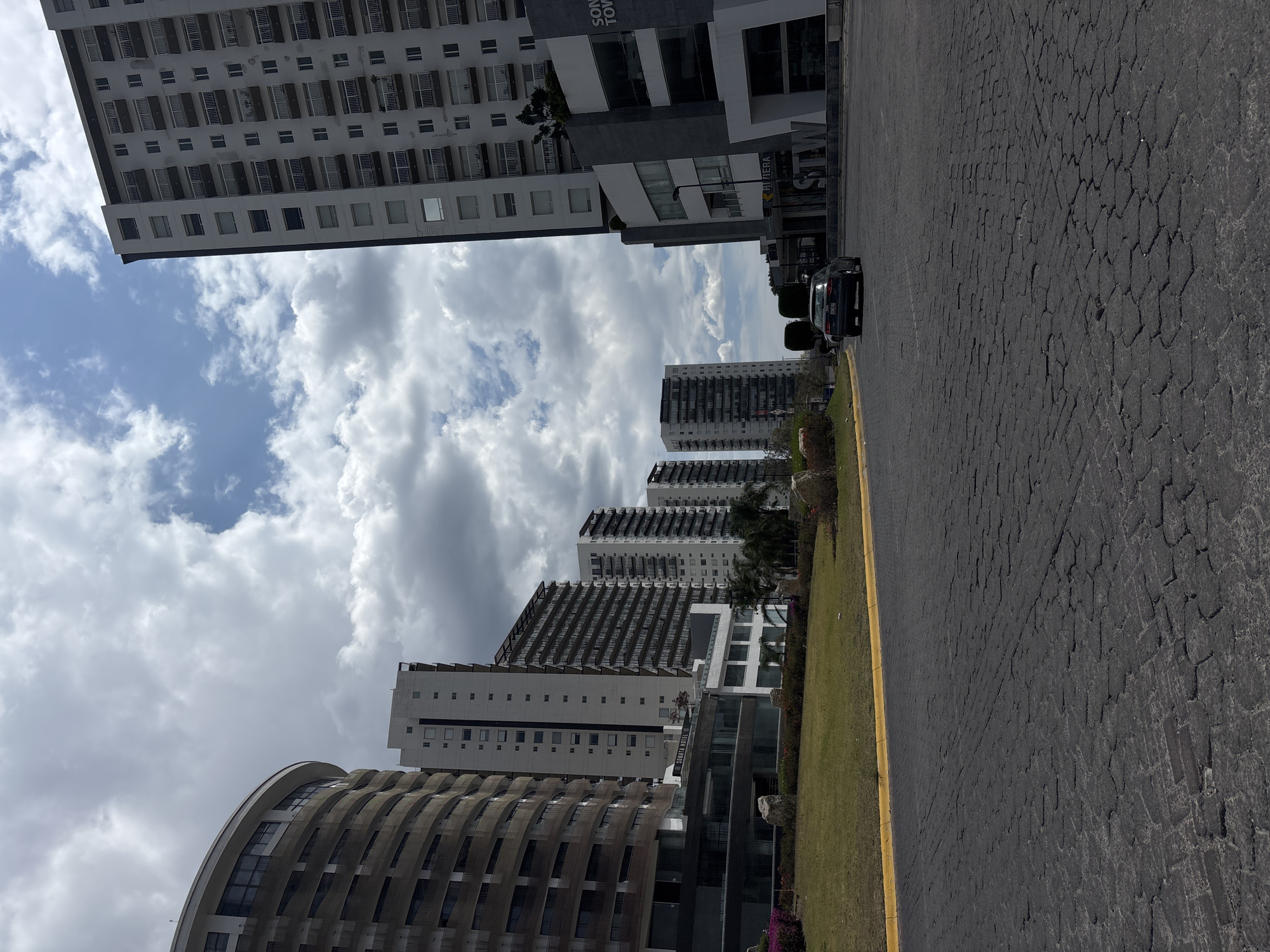 The image depicts a street scene in San Bernardino Tlaxcalancingo, Mexico. Several tall, modern buildings are visible against a cloudy sky. The building closest to the viewer has a distinctive curved facade with horizontal bands. Other buildings in the background are rectangular with balconies. A black car is parked along the right side of the road, which is made of dark paving stones. A strip of grass and some shrubbery separates the road from the buildings. A yellow line is painted along the edge of the road.