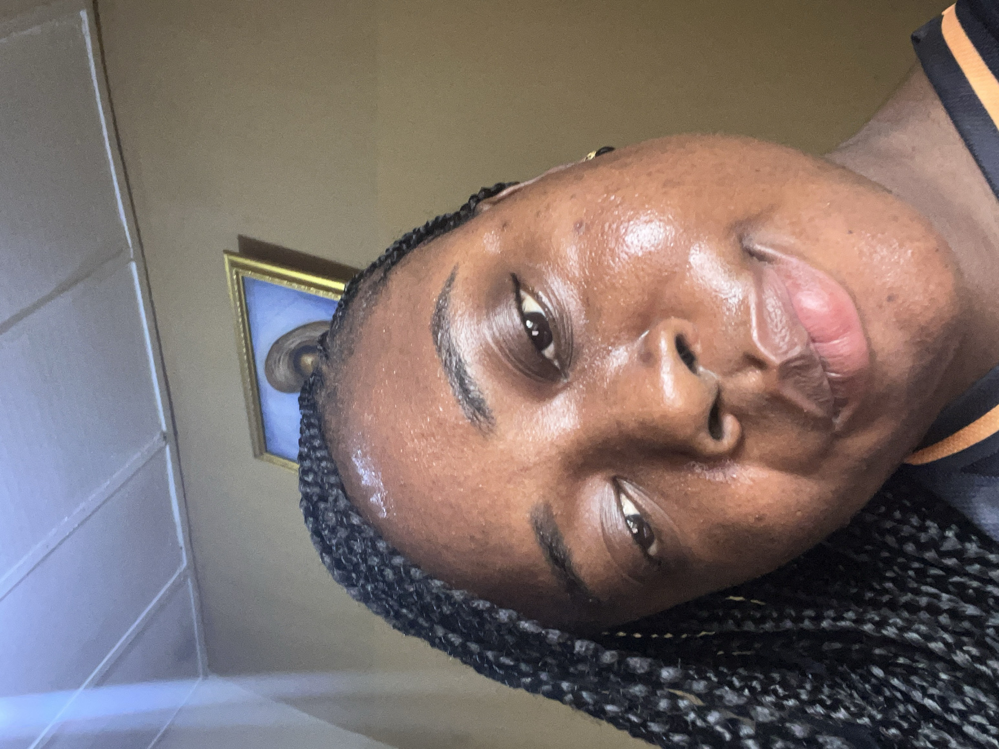 A close-up, angled portrait of a dark-skinned individual with braided hair. The individual's face is centered in the frame, with the eye on the lower right appearing prominent. The setting appears to be indoors, based on the wall and framed picture in the background. There's no specific element visible in the image to suggest the precise location in Madalla, Nigeria.