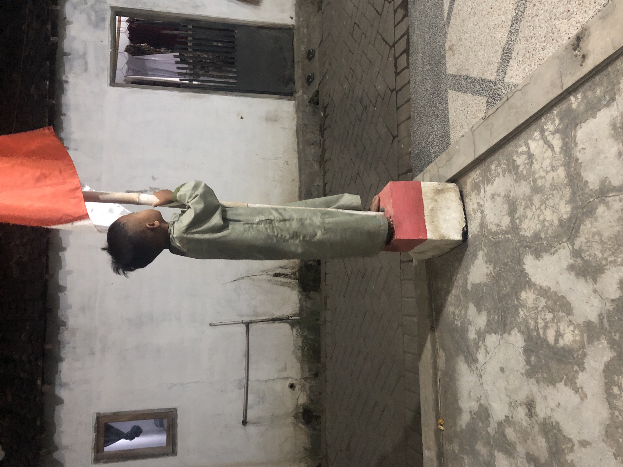 A young child stands on a red and white concrete block, holding a bamboo pole with an Indonesian flag. The child is wearing a long, light green robe. The scene appears to be in a residential area, with a white building in the background that has a wooden door with vertical bars and a small window. The ground is paved with brick and concrete. The image is taken at night or in low light conditions.