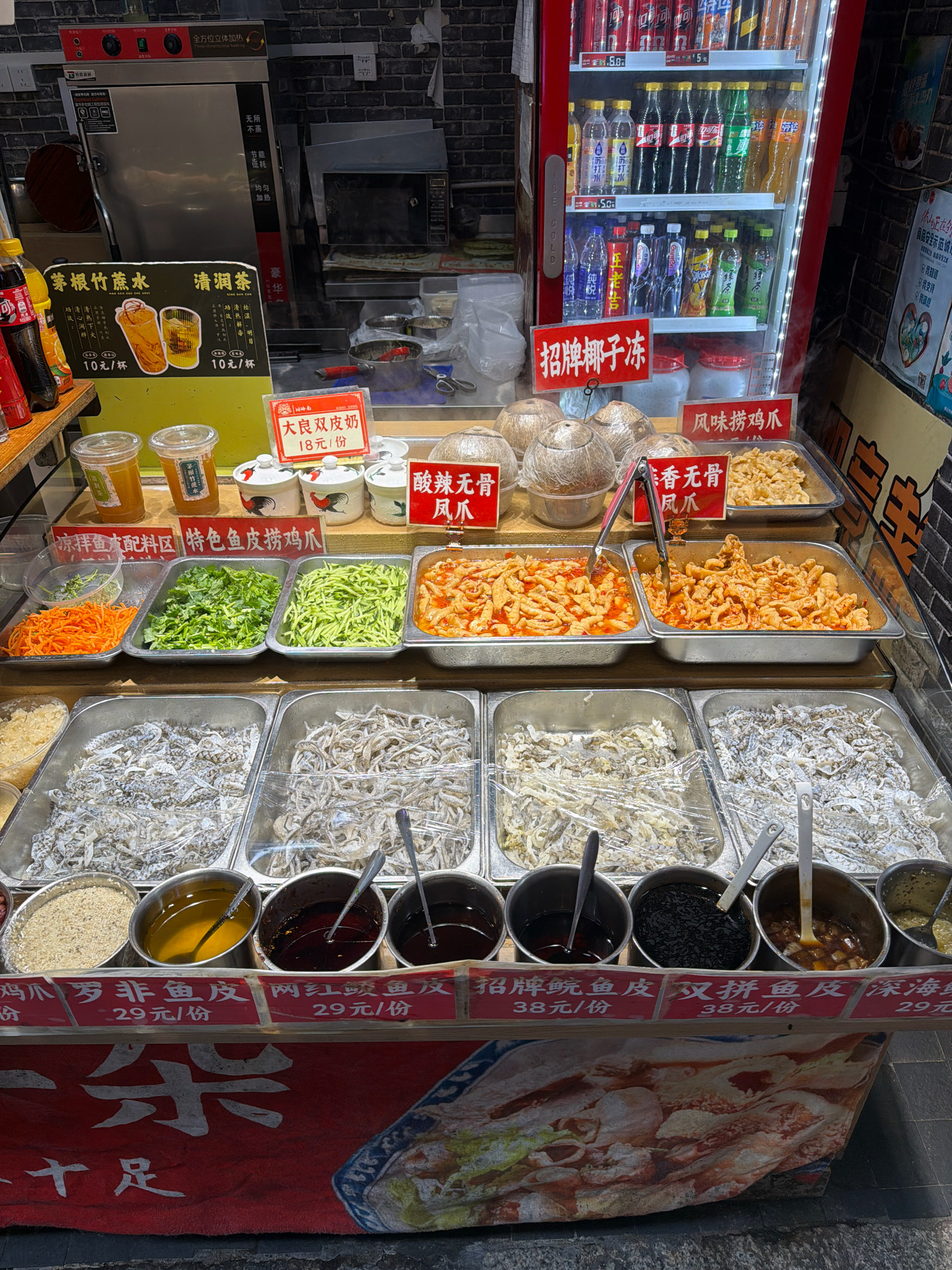 The image depicts a street food stall in Chancheng District, China. The stall features a refrigerated display case stocked with various bottled beverages, including Coca-Cola, Sprite, and other unnamed drinks. In front of the refrigerator, a variety of prepared food items are displayed in stainless steel trays. These include what appear to be pickled vegetables such as shredded carrots and cucumbers, as well as dishes of seasoned chicken feet and small fried fish or seafood. Several small bowls containing different dipping sauces or condiments are also visible. In the background, a large stainless steel steamer and a microwave oven are present, suggesting food preparation is done on-site. Signs in Chinese are displayed throughout the stall, indicating prices and product names. The overall environment suggests a busy urban setting.