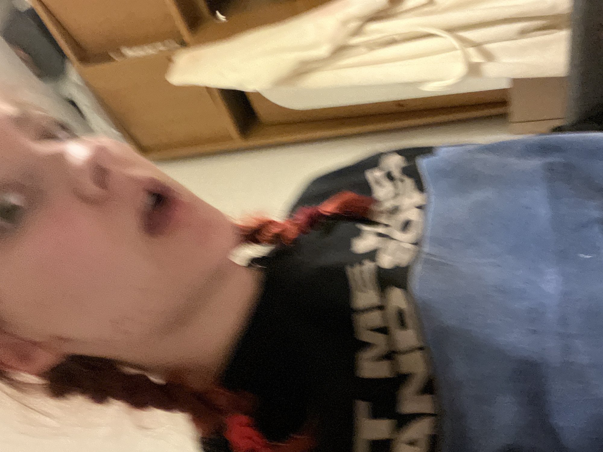 A blurry, close-up photograph shows a person with pale skin, open mouth, and green eyes. They appear to be wearing a black shirt with white lettering and a blue garment. In the background, a wooden shelf and a white cloth with a drawstring are visible. The image lacks identifiable details that would indicate a location in Sofia, Bulgaria.