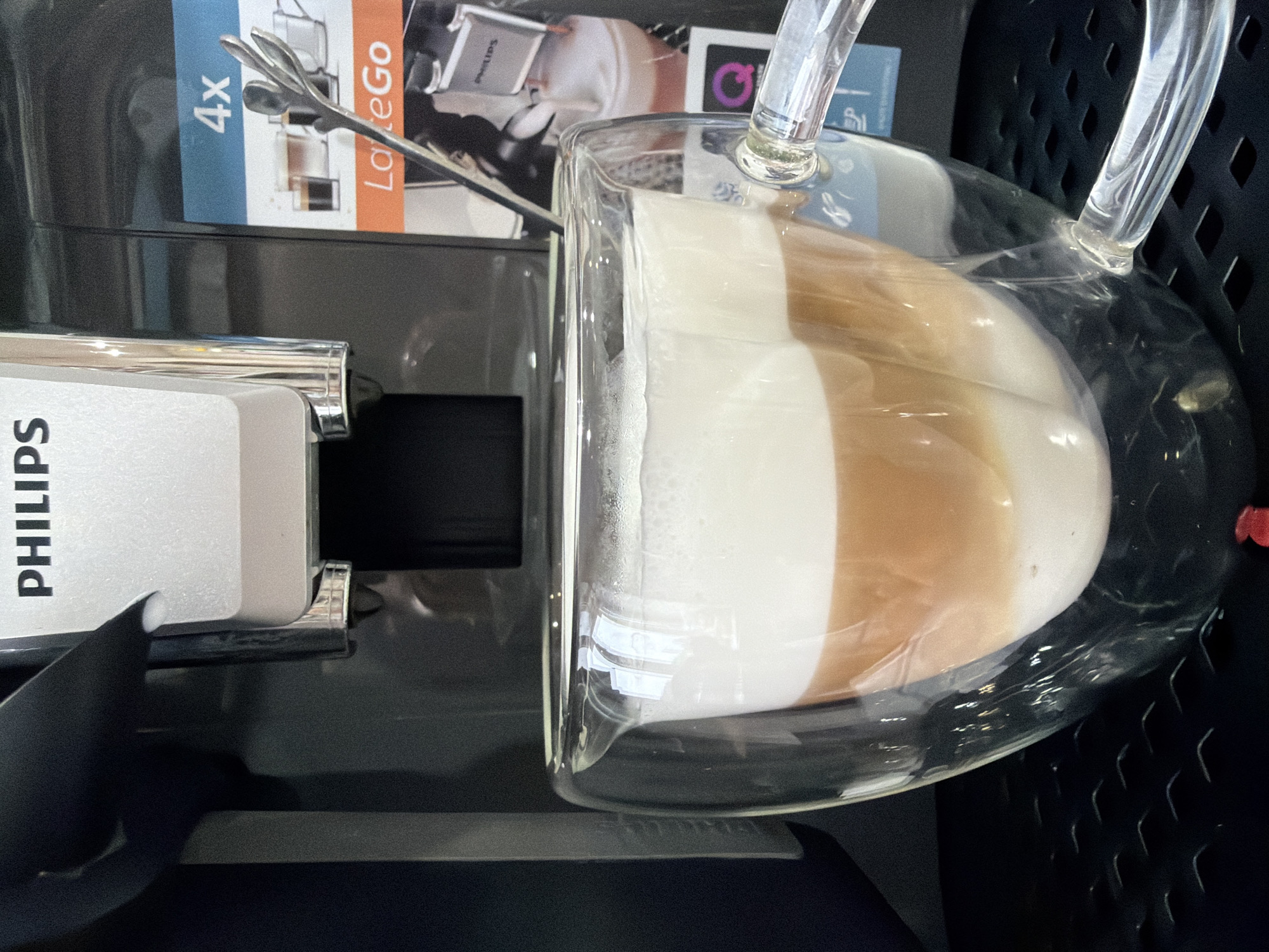 The image shows a Philips LatteGo coffee machine dispensing a latte into a clear glass mug. The mug has a layered drink with a white foam layer at the bottom and a brown coffee layer on top. The coffee machine is black and has a silver Philips logo on the front. In the background, there is a box with "LatteGo" written on it. The location is not discernible from the image.