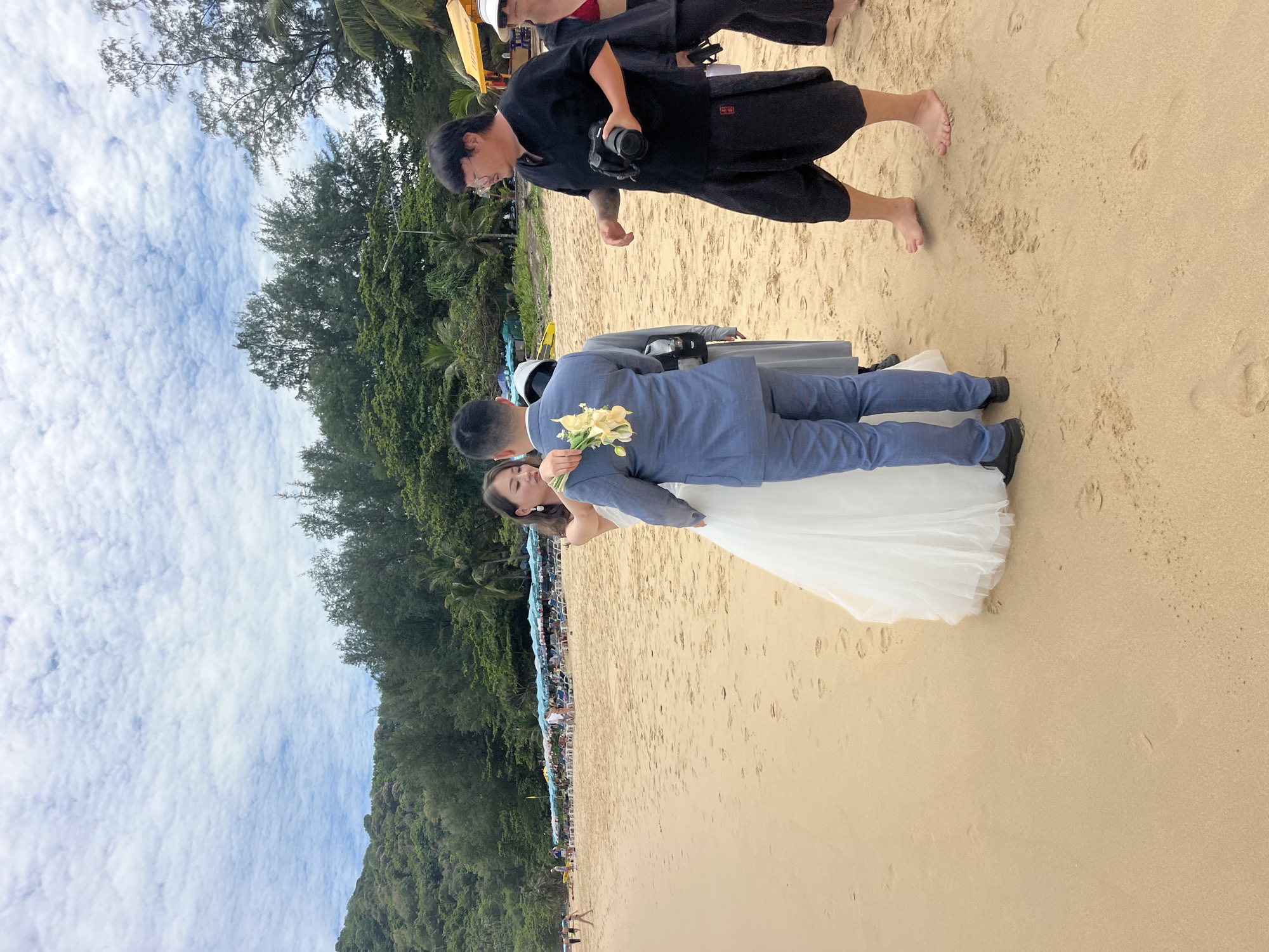 On a sandy beach in Rawai, Thailand, a wedding photoshoot is underway. A bride in a white wedding dress and a groom in a blue suit stand facing each other. The bride is holding a bouquet of white lilies. A photographer, dressed in black, stands to the right of the couple, holding a camera. A woman in a red bikini top and a black cover-up stands behind the photographer. In the background, beach umbrellas and lounge chairs are visible, along with lush green hills and palm trees. The sky is partly cloudy.