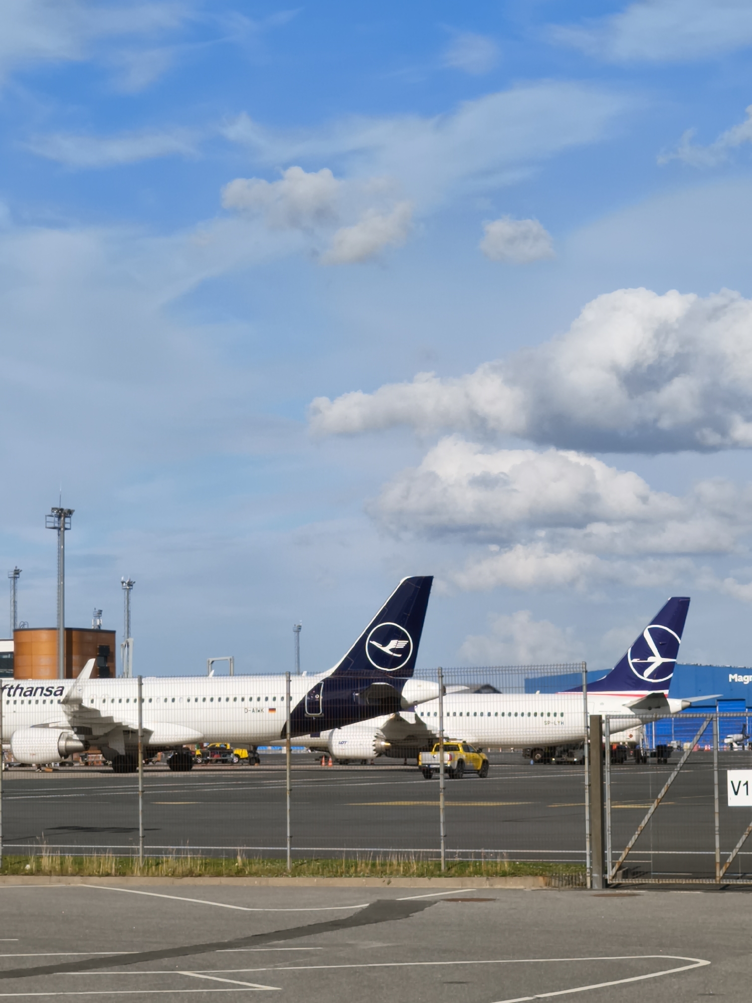 The image shows a daytime exterior view of an airport tarmac at Tallinn Airport, Estonia. Two commercial passenger jets are parked side-by-side, with a chain-link fence in the foreground. The jet on the left has "Lufthansa" branding visible on its fuselage and a Lufthansa logo on its tail. The jet on the right has a different airline logo on its tail, appearing to be LOT Polish Airlines. The sky is blue with scattered white clouds. In the background, there are airport structures, including a building with an orange facade, and utility towers with lights. A yellow service vehicle is parked between the two aircraft. The ground is asphalt, marked with white lines. The overall scene depicts a typical operational environment at a major airport.