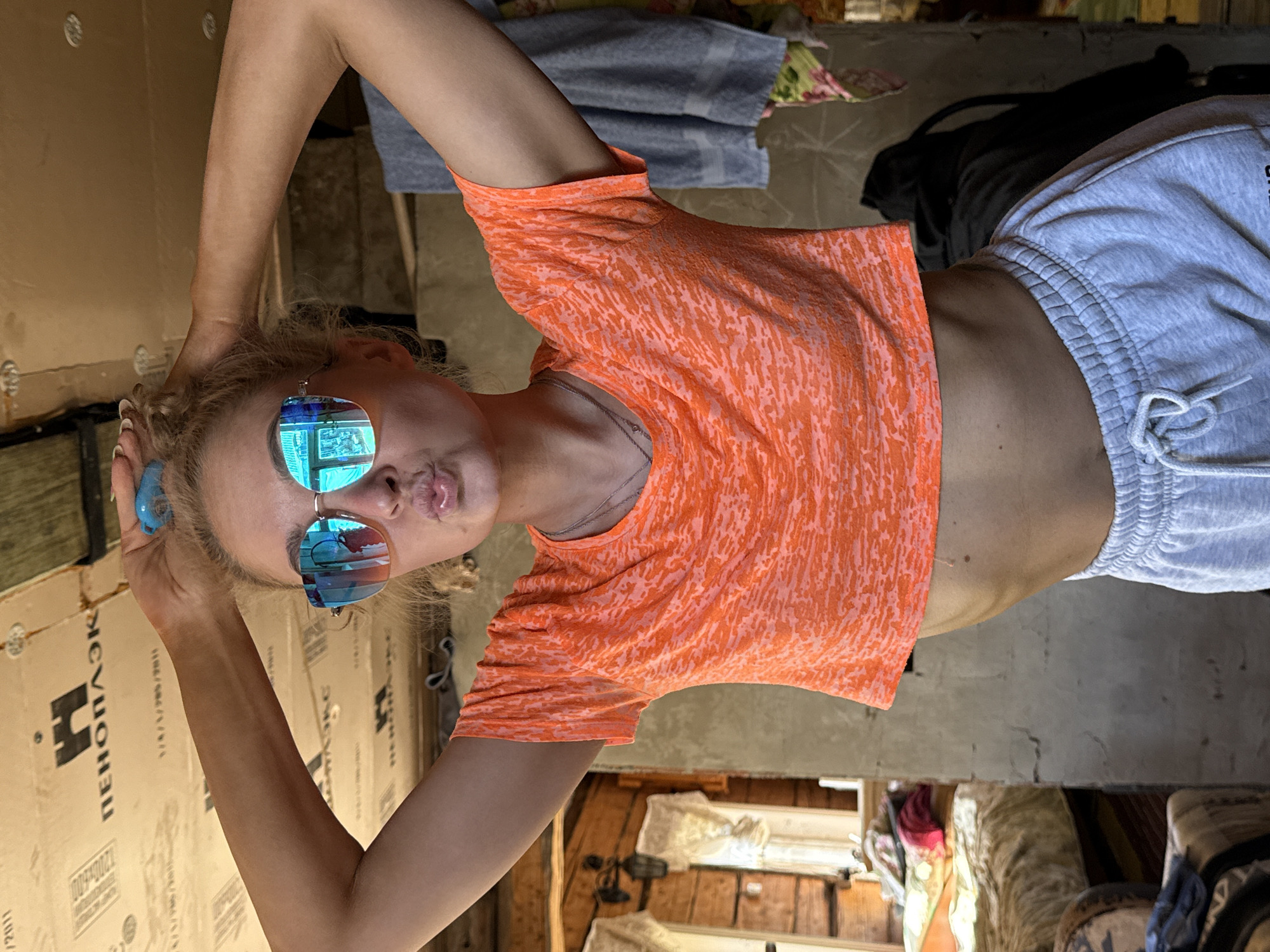 A young woman is posing for a photo. She is wearing sunglasses and an orange shirt with gray shorts. She has her hands raised above her head. The surroundings appear to be a room under construction or renovation, with unfinished walls and various objects scattered around. It is not possible to determine the location in Rosstani, Russia from the image.