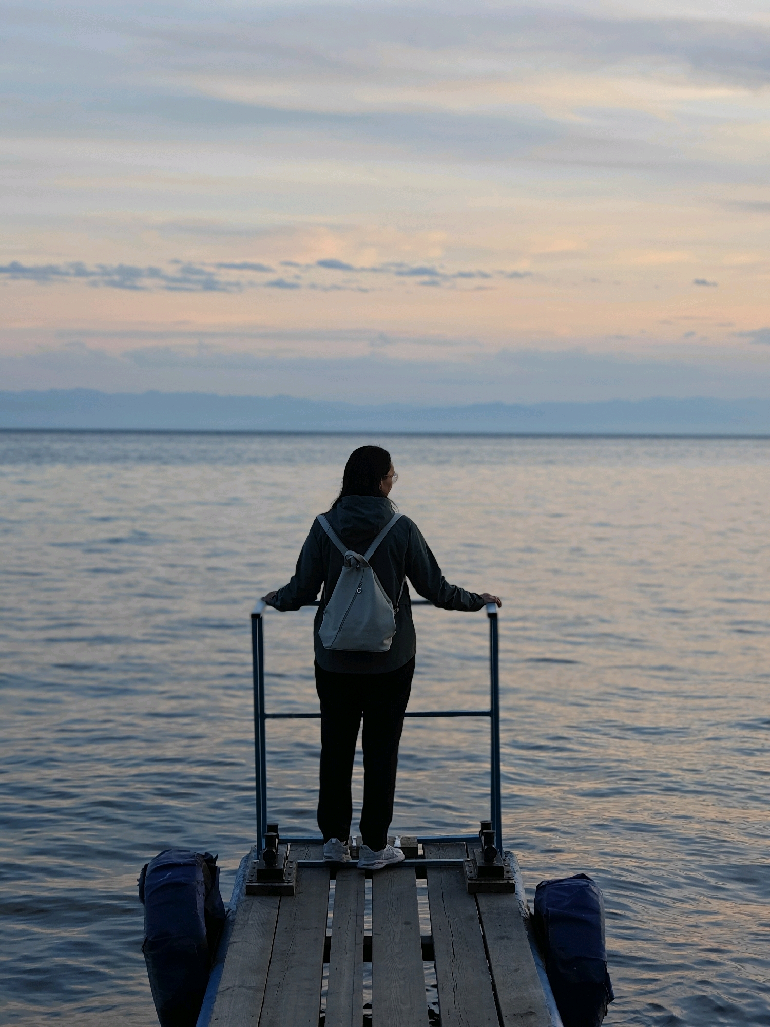 A woman stands on a wooden dock, facing away from the camera, looking out at a large body of water. The water is calm and reflects the soft colors of the sky, which is a mix of pale blue, pink, and orange, suggesting it is either sunrise or sunset. In the distance, faint, hazy mountains can be seen on the horizon. The woman is wearing a dark hooded jacket, dark pants, and light-colored sneakers. She has a light gray backpack on her shoulders and is holding onto a metal railing. The dock is made of weathered wooden planks and has blue inflatable fenders attached to its sides. The scene appears serene and tranquil, possibly capturing a moment of reflection in nature. The location is the city of Listvyanka, Russia, known for its proximity to Lake Baikal.