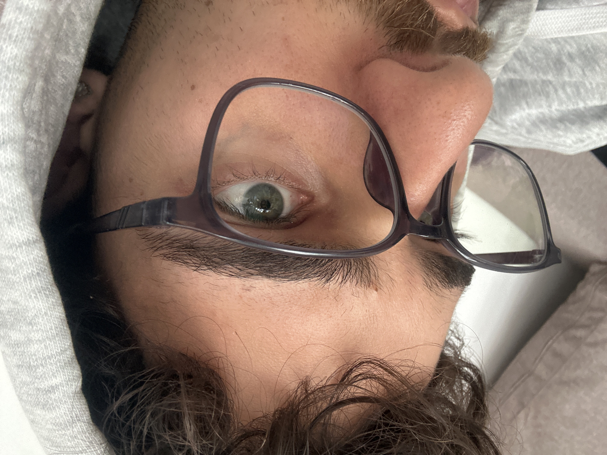 This close-up portrait shows a person's face, oriented sideways, with their head tilted 90 degrees counter-clockwise. The main subject is an eye, which has a distinct green-hazel iris and is looking directly at the viewer. The person is wearing dark-framed glasses, with one lens covering part of the eye and the bridge resting on the nose.
Visible details include thick, dark eyebrows and dark, wavy hair extending from the forehead area. Above the nose, there's light brown stubble or a mustache. The person is wearing a light grey hooded sweatshirt, with the fabric visible on the left side of the frame and around the head. The skin shows visible pores and some minor blemishes. Reflections of a light source are present in both the glasses and the pupil of the eye. The image captures a direct, neutral gaze. There is no visible text in the image. The location context for this image is Troyan, Bulgaria.