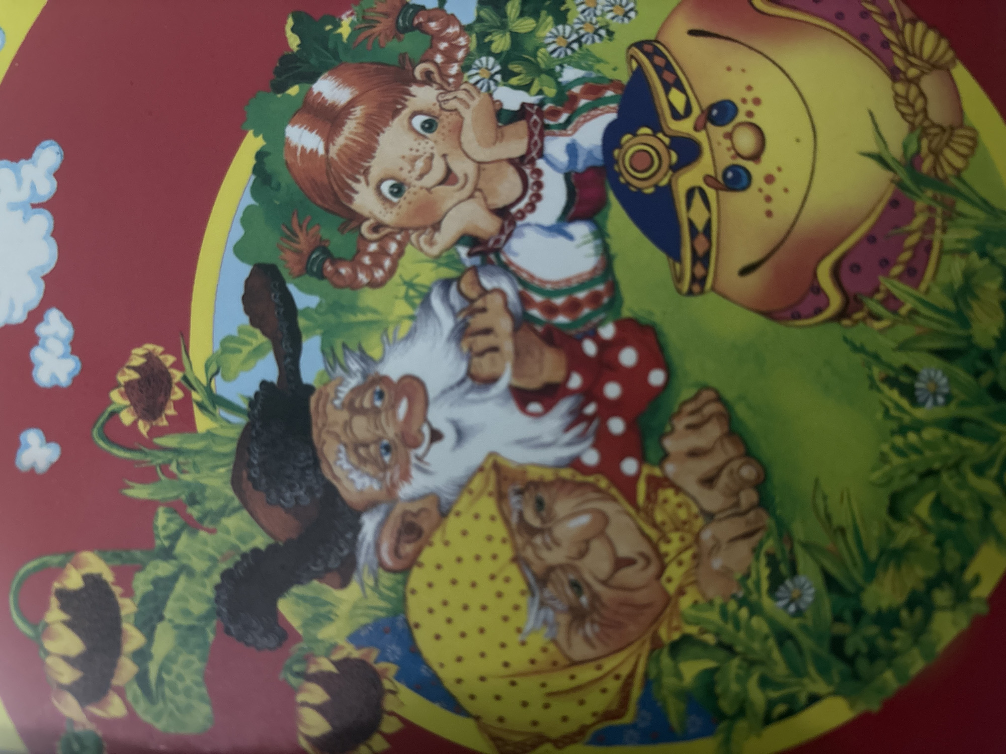 This is an image of a colorful illustration featuring several cartoon characters, likely from a children's story or folk tale. The subjects include a young girl with red braided pigtails, freckles, and a mischievous grin. She is wearing a traditional-style dress with red and green embroidery. Beside her are two older individuals, a man and a woman, who appear to be gnomes or dwarves. The man has a long white beard, is wearing a dark hat and a red polka-dot shirt, and has a large, jovial smile. The woman is wearing a yellow polka-dot headscarf and also has a warm, grandmotherly expression. They are all situated in a grassy, outdoor setting with sunflowers and small white flowers visible. The overall mood is cheerful and whimsical. To the right of the scene is a large, decorative object, possibly a pot or a shield, adorned with blue and yellow patterns. The image appears to be a painting or drawing with a slightly rustic, storybook aesthetic. There is no visible text in the image. Given the provided location context of a rural settlement in Russia, the imagery might be inspired by Slavic folklore or fairy tales. The time of day and weather are not explicitly indicated but the bright lighting suggests daytime and fair weather.