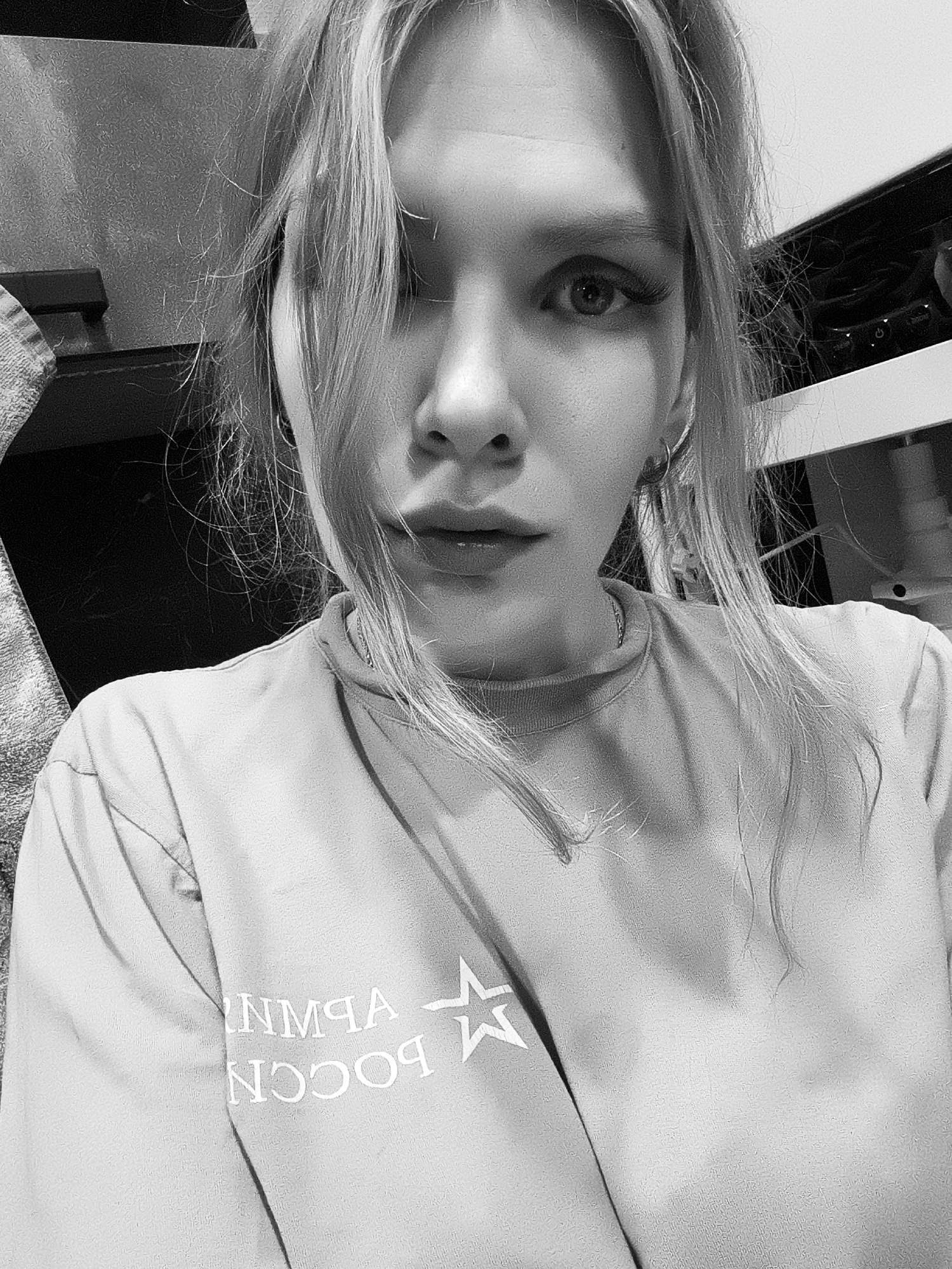 A black and white selfie taken by a woman in a room that appears to be her home, evidenced by personal items in the background. The woman has blonde hair, styled with some strands covering part of her face. She is wearing a t-shirt with text written in Russian, likely indicating a location or group affiliation. The text visible on the shirt reads "ВОССОЙ" and "ИМЧА," likely part of a larger phrase. The logo of a star is present next to the text. The background is out of focus, but shows a combination of light and dark objects including what appears to be a towel to the left. The setting is unknown and location identification cannot be confirmed with current information.