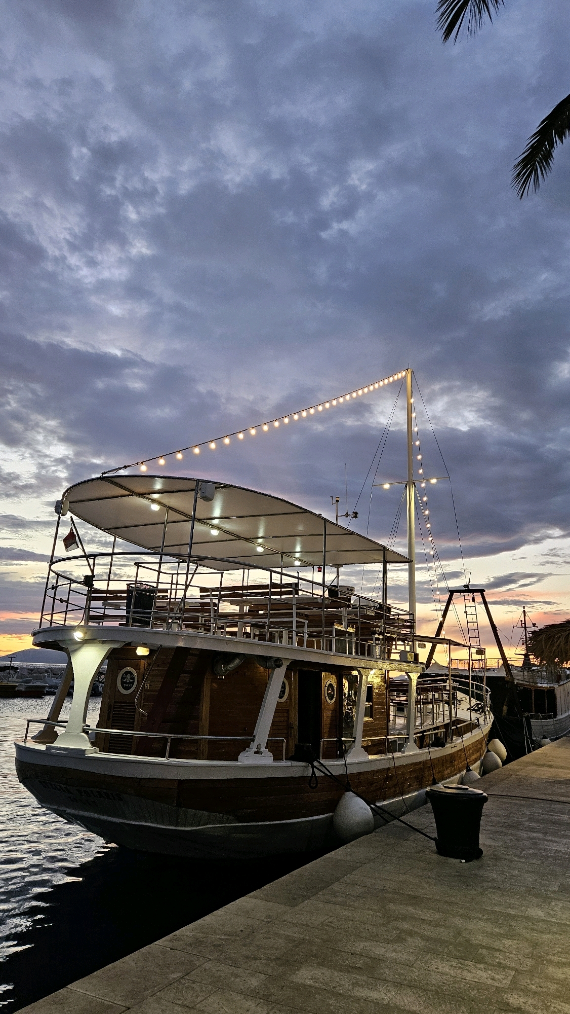 The image depicts a moored wooden boat at dusk in the city of Općina Tučepi, Croatia. The boat, illuminated by strings of fairy lights and interior lighting, features multiple decks with seating arrangements. It is docked alongside a concrete pier, with the calm water of the harbor visible to the left. The sky is filled with dramatic, cloudy twilight colors, transitioning from warm oranges and pinks near the horizon to darker blues and grays overhead. A palm frond is visible in the upper right corner, indicating a coastal or tropical setting. The overall atmosphere is serene, capturing the boat at rest by the water as day transitions into night.