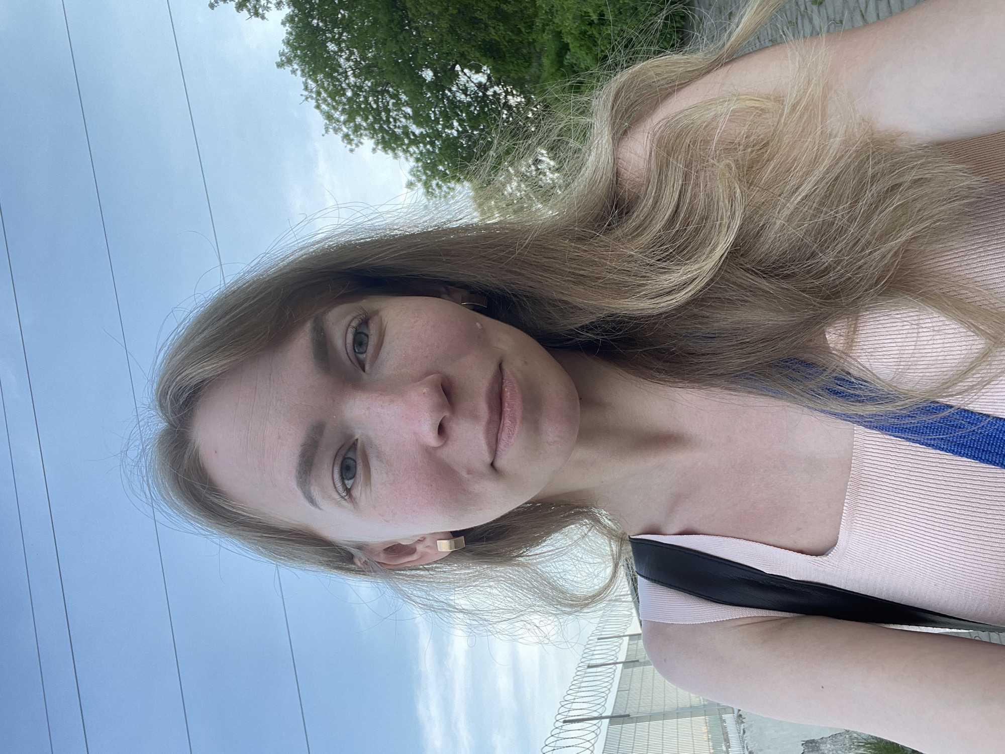 A blonde woman stands outdoors, facing the camera, with a backdrop of the sky, trees, power lines, and a building with fencing in the distance. She wears a light pink top with blue and black straps and a gold earring. The location may be in the city of Perm, Russia.