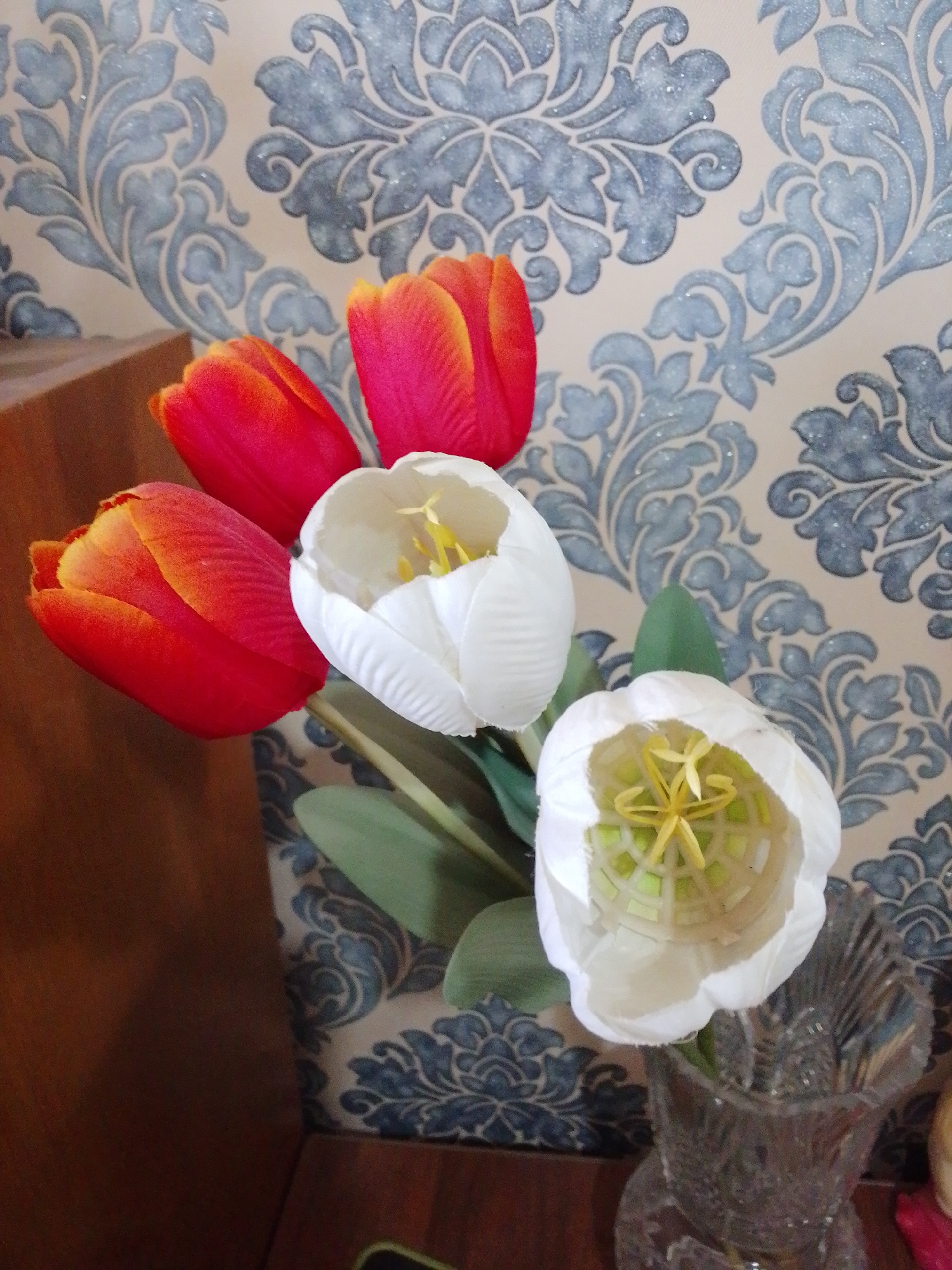 A close-up, eye-level shot captures a bouquet of five tulips, two white and three red with orange tips, arranged in a clear glass vase. The vase sits on a dark brown surface, possibly a wooden table, with a patterned wallpaper featuring light beige and blue damask designs serving as the backdrop. The lighting is moderate, casting soft shadows and highlighting the textures of the flowers and wallpaper. The bouquet is positioned to the right of center, with the red tulips in the foreground and the white tulips slightly behind and to the left. The overall scene is static and appears to be an indoor still life.